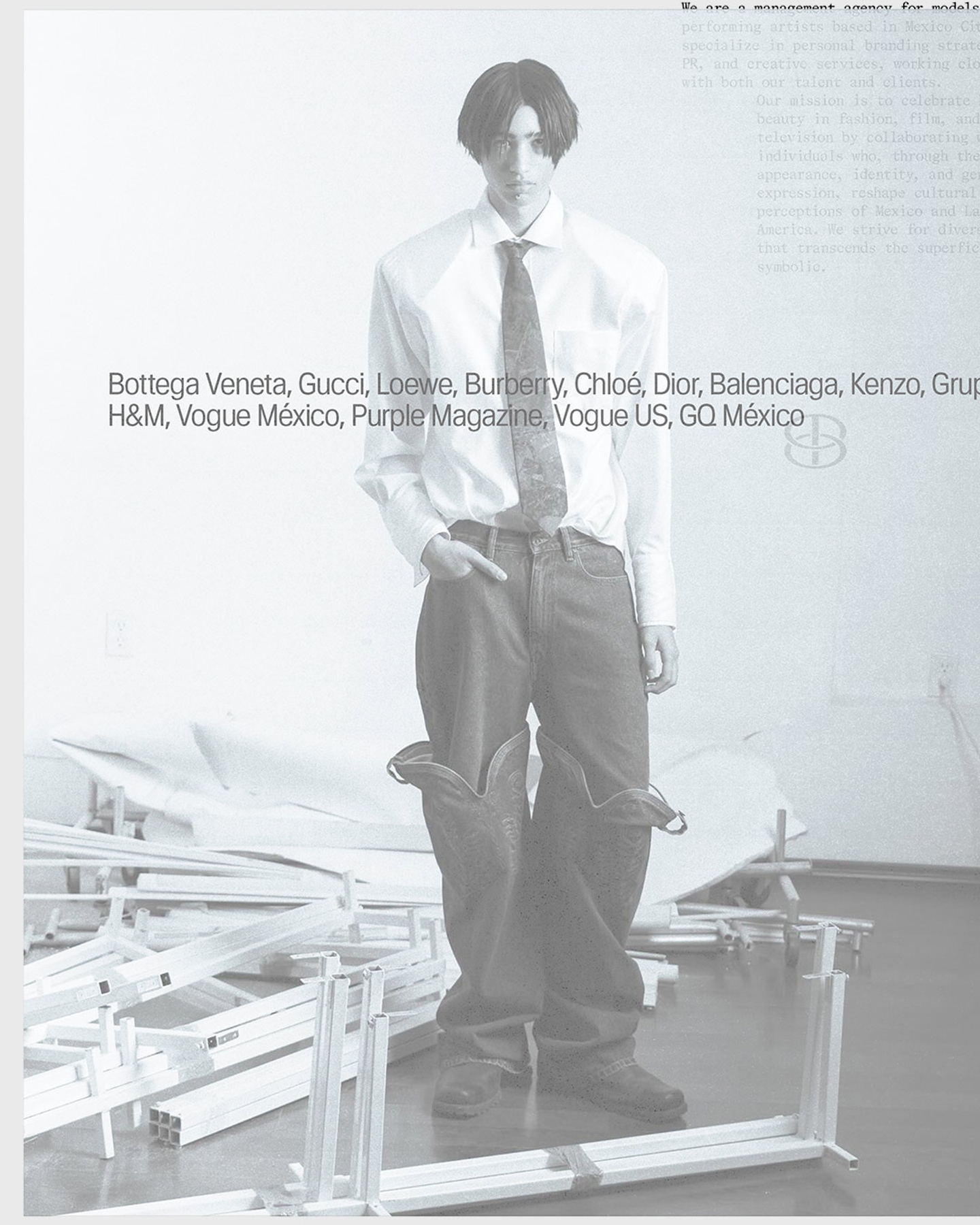 Black-and-white fashion editorial photograph of a male model wearing a white dress shirt, patterned tie, and oversized denim jeans with leather boots, standing on an industrial platform surrounded by white metal scaffolding.