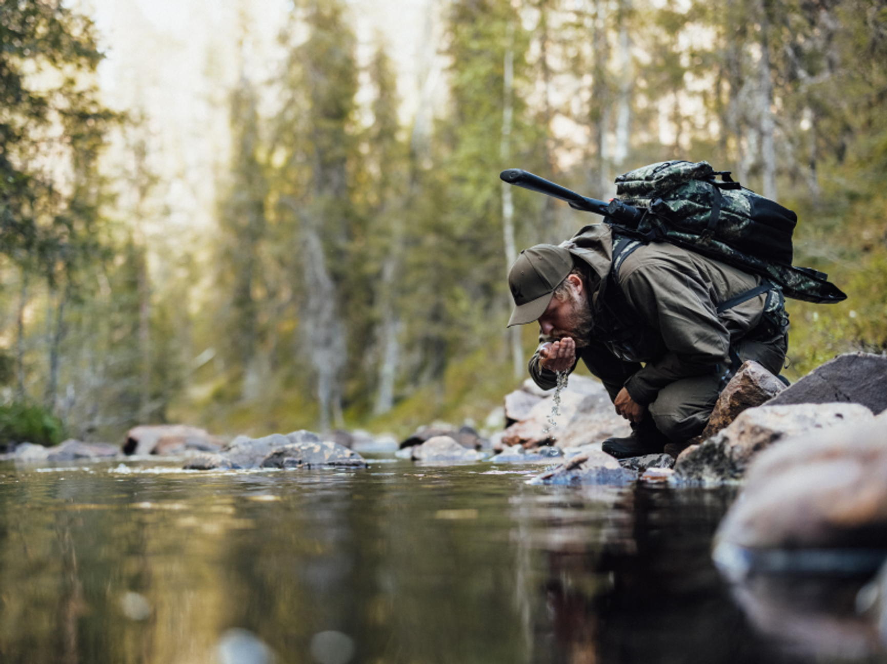 man med vapenryggsäck som dricker ur en skogsbäck