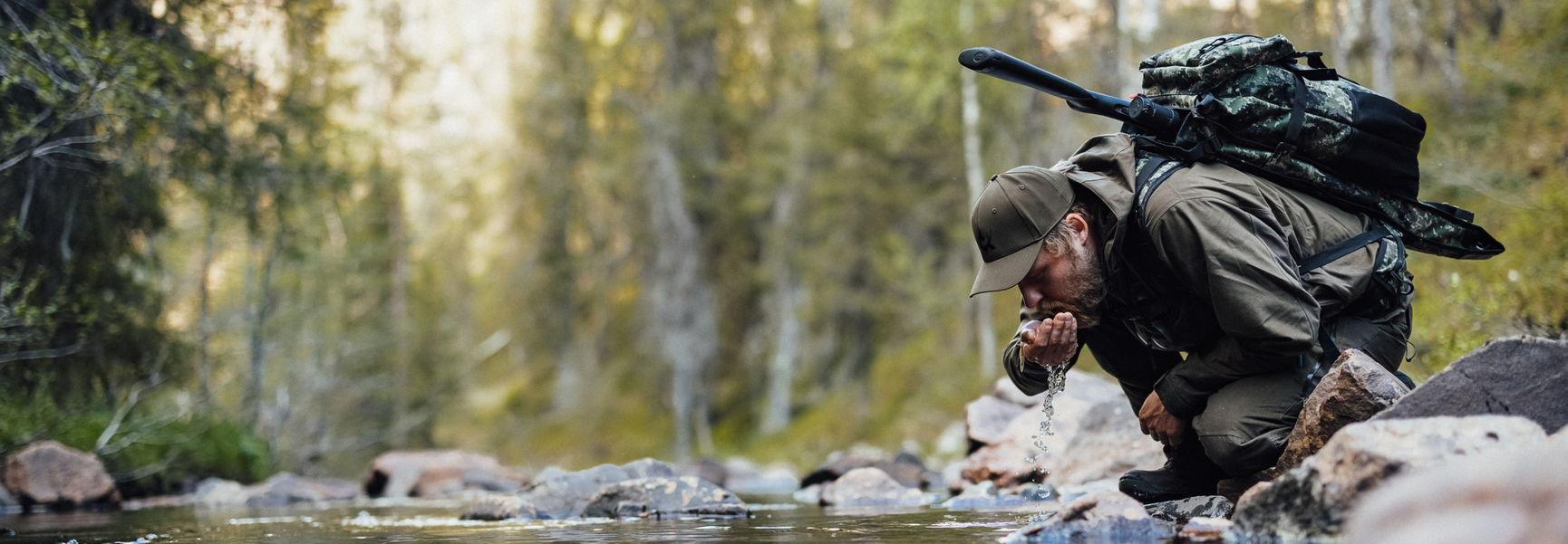 man med vapenryggsäck som dricker ur en skogsbäck