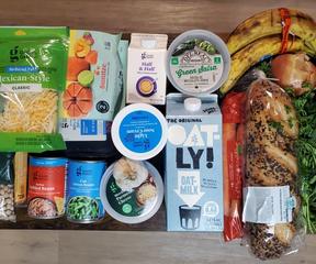 Groceries spead out on a table including oat milk, a loaf of bread, cans, cheese, and other items.