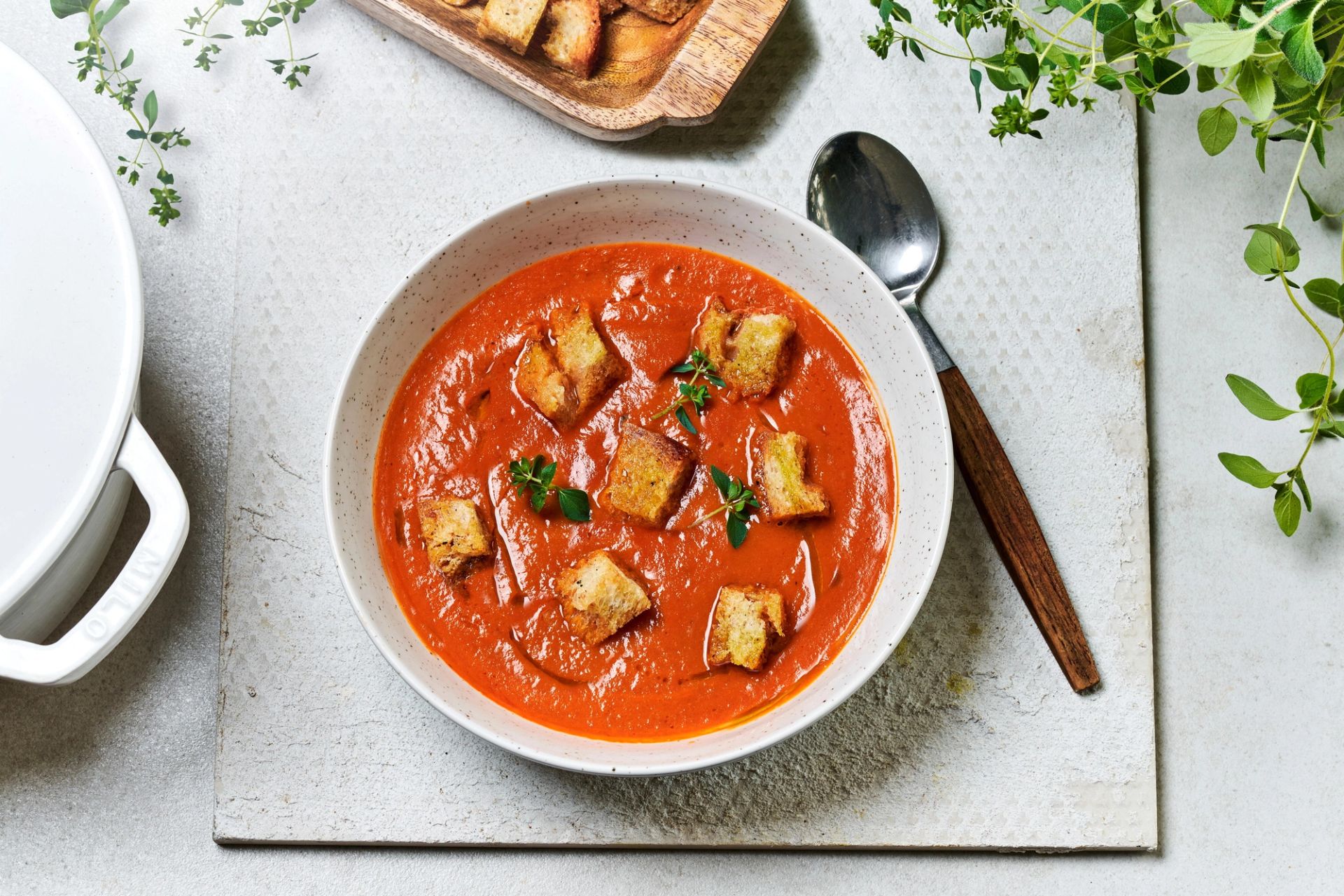 To hvite boller fylt med kremet tomatsuppe, toppet med brødkrutonger, friske basilikumblader og en tynn ring av olje. Bollene står på et trebord, omgitt av hele tomater, en skje og en liten skål med pepperkorn.