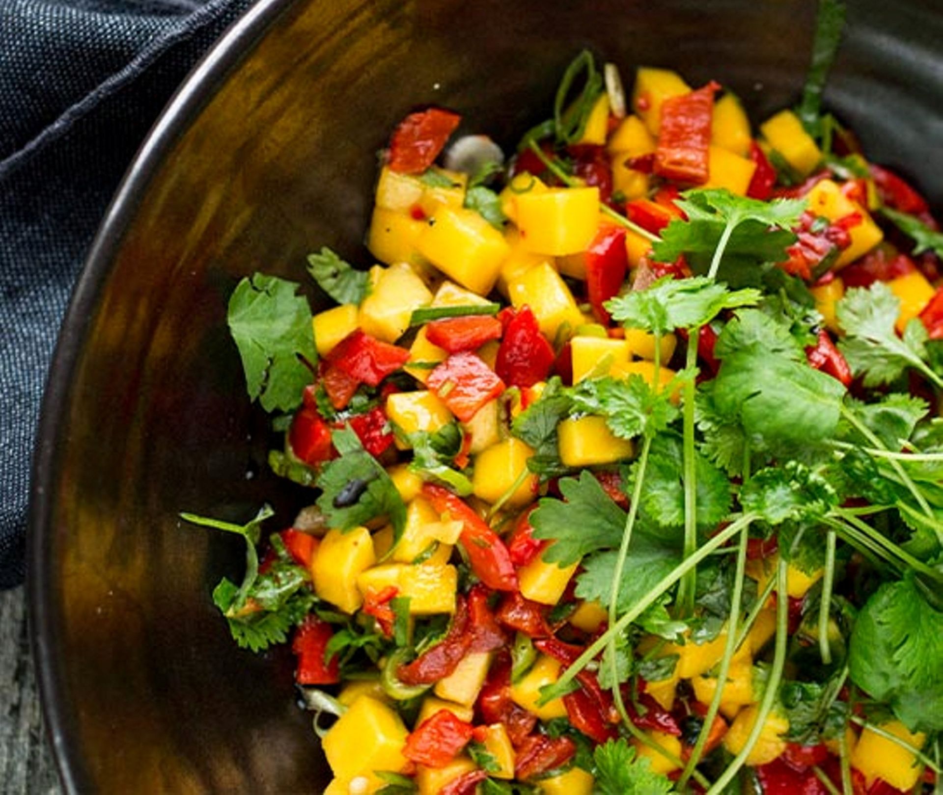 En svart skål fylt med fargerik mango‑ og paprikasalsa. De gule mangobitene og røde paprikaene er blandet med finhakket grønn urt, og frisk koriander ligger på toppen. En mørk stoffserviett ligger ved siden av skålen.