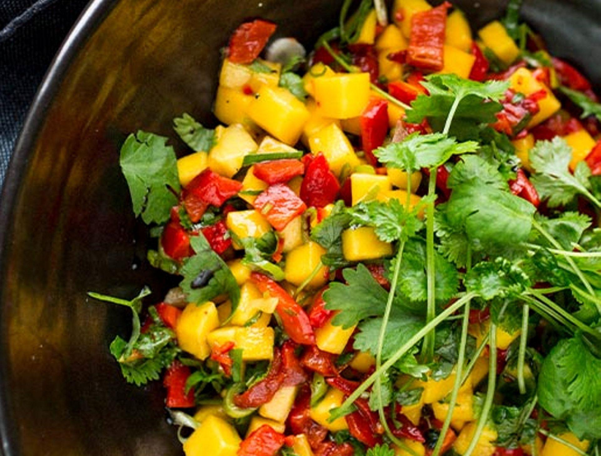 En svart skål fylt med fargerik mango‑ og paprikasalsa. De gule mangobitene og røde paprikaene er blandet med finhakket grønn urt, og frisk koriander ligger på toppen. En mørk stoffserviett ligger ved siden av skålen.