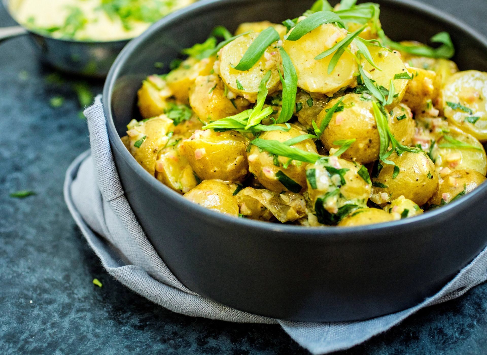 Potetsalat med urter og sennepsdressing toppet med vårløk, servert i en mørk grå skål på et grått kjøkkenhåndkle.