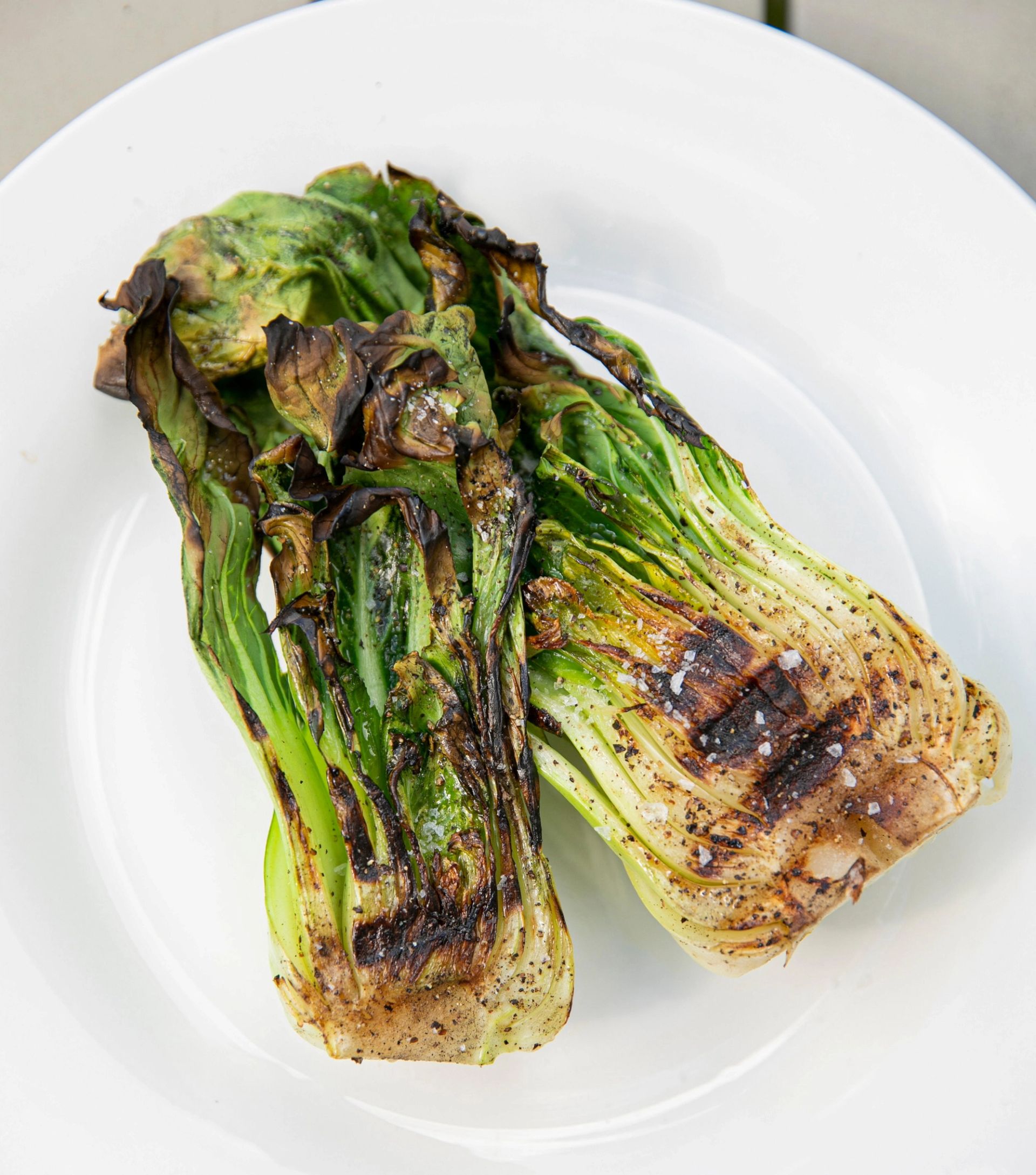 To halve pak choy som er grillet, lagt på en hvit tallerken. Grønnsaken har tydelige grillmerker, lett forkullede bladkanter og er krydret med salt og pepper. Fargene varierer fra lys grønn i stilkene til mørkere, brungrillede partier på bladene.