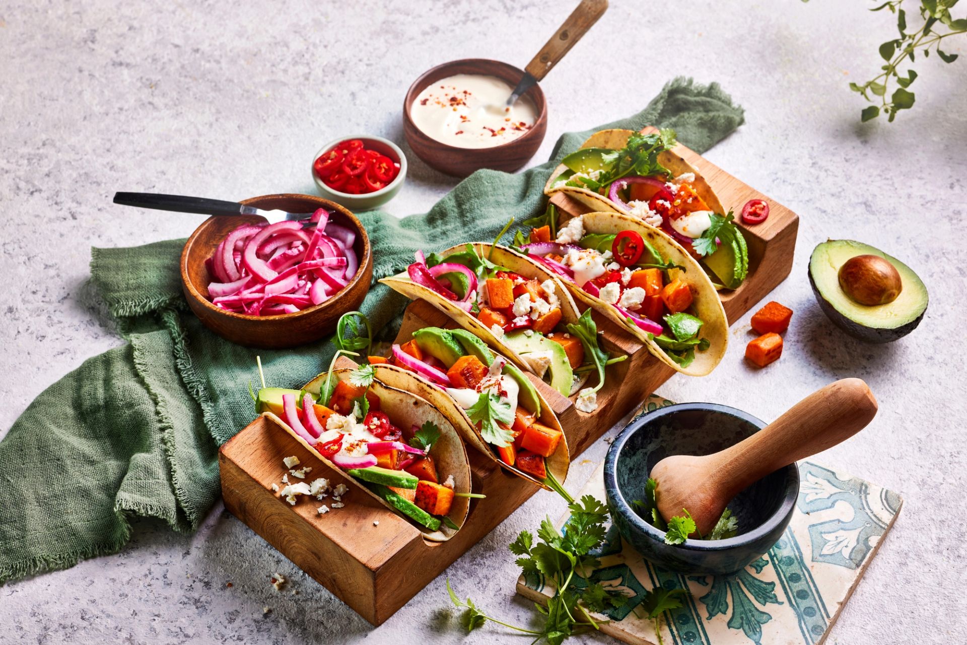 Oversiktsbilde av fire tacos fylt med ovnsbakte terninger av potet, avokadoskiver, syltet rødløk, frisk koriander og smuldret ost, toppet med saus og skiver av chili.