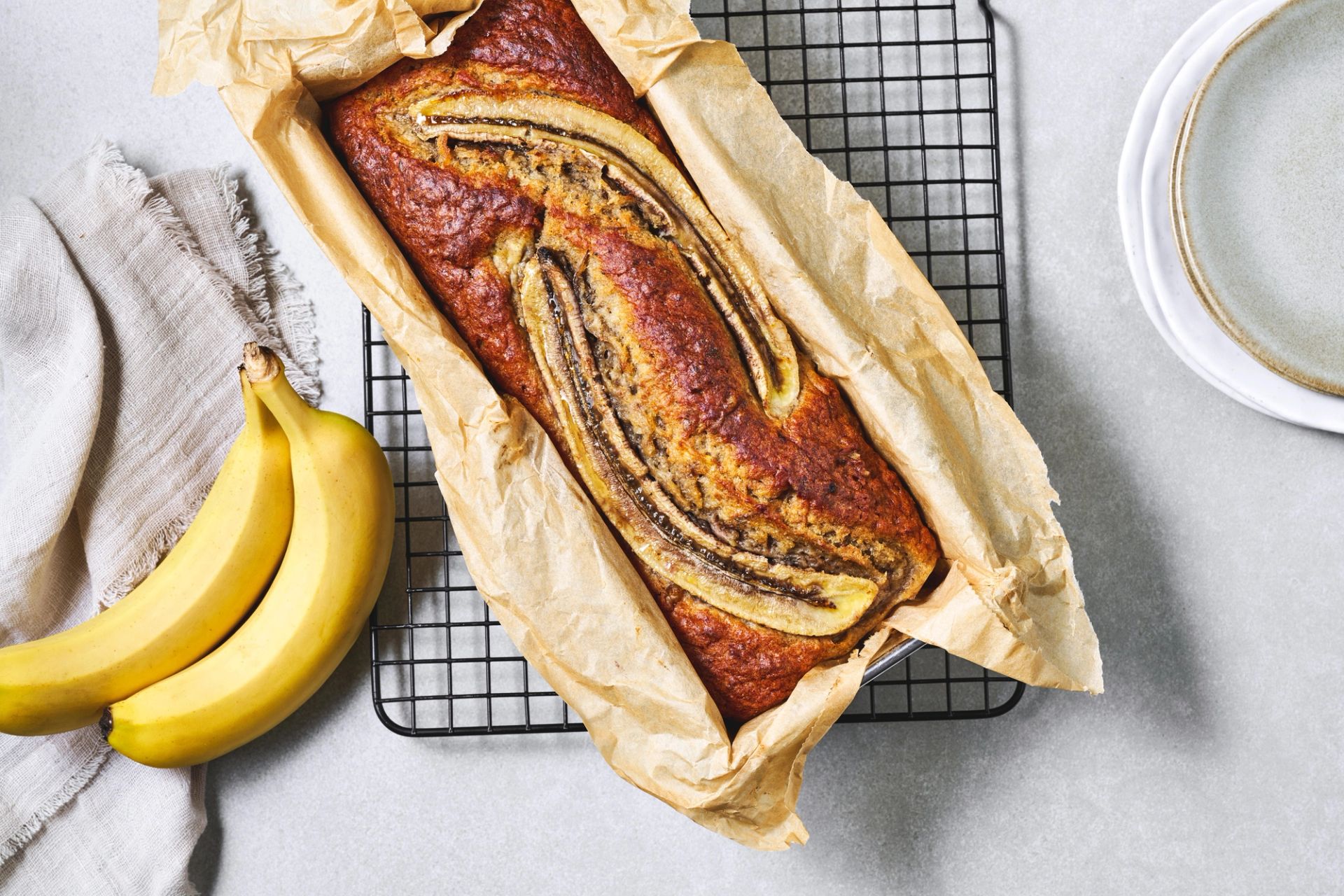  Hjemmelaget bananbrød på et fat, med flere skiver skåret opp, dekorert med bananskiver, friske bananer ved siden av og et glass melk i bakgrunnen.