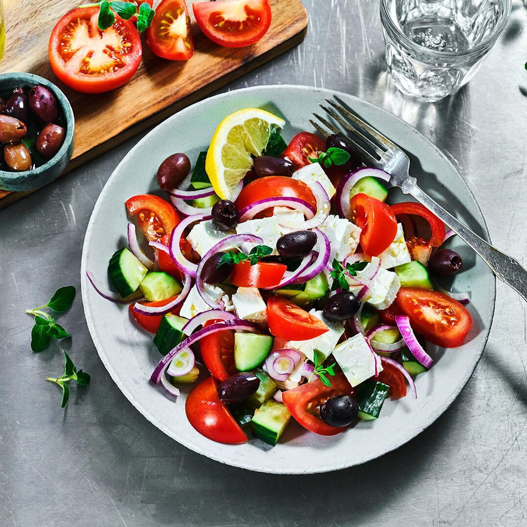 Gresk salat i en skål. trefjøl ved siden av med kuttet tomat og oliven.