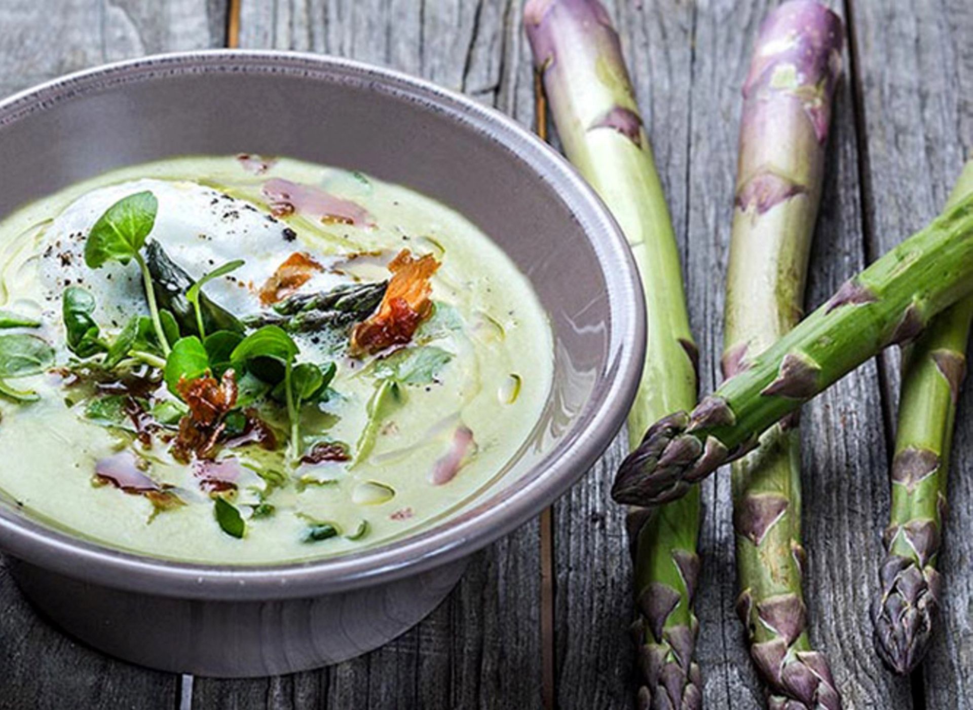 Grønn aspargessuppe i en grå skål, toppet med rømme, ruccolaskudd og sprøstekt bacon, servert ved siden av friske asparges på et trebord.