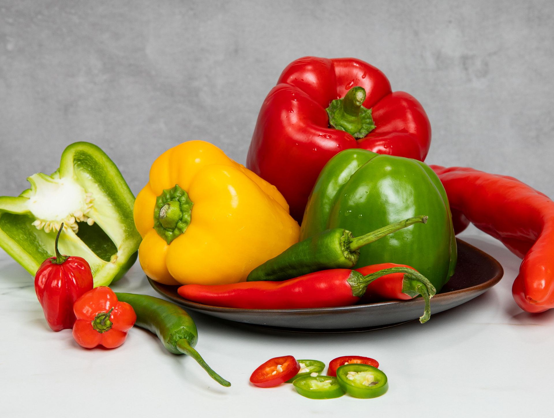Et fat med fargerike paprika og chili på et hvitt bord: rød, gul og grønn paprika, lange røde chilipepper, grønne chilipepper og noen oppskårne chiliskiver, mot en grå bakgrunn.