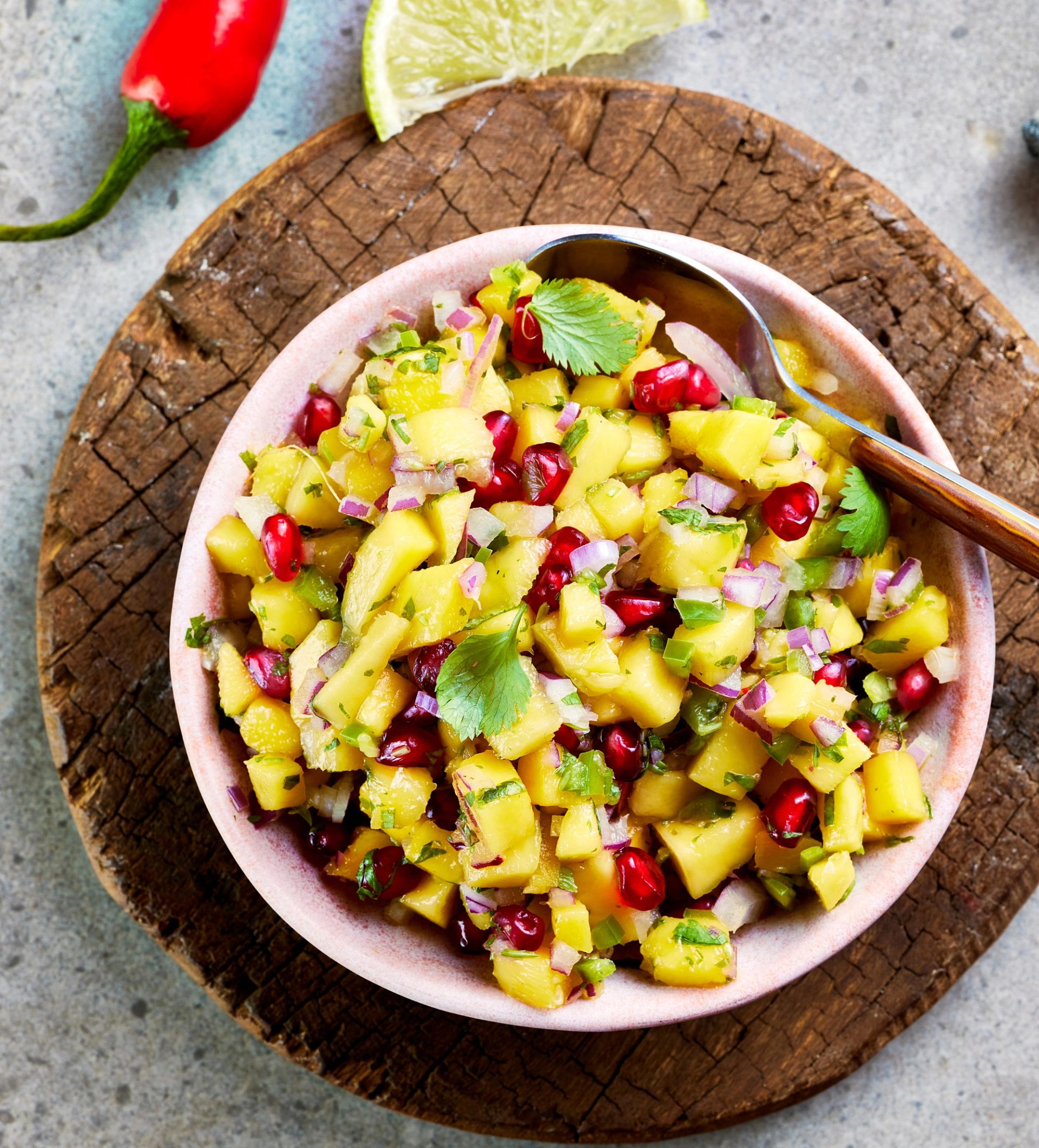 En bolle med frisk mangosalsa laget av terninger av mango, granateplekjerner, finhakket rødløk og urter, servert med en skje og plassert på et rundt trefat, med chili og lime i bakgrunnen.