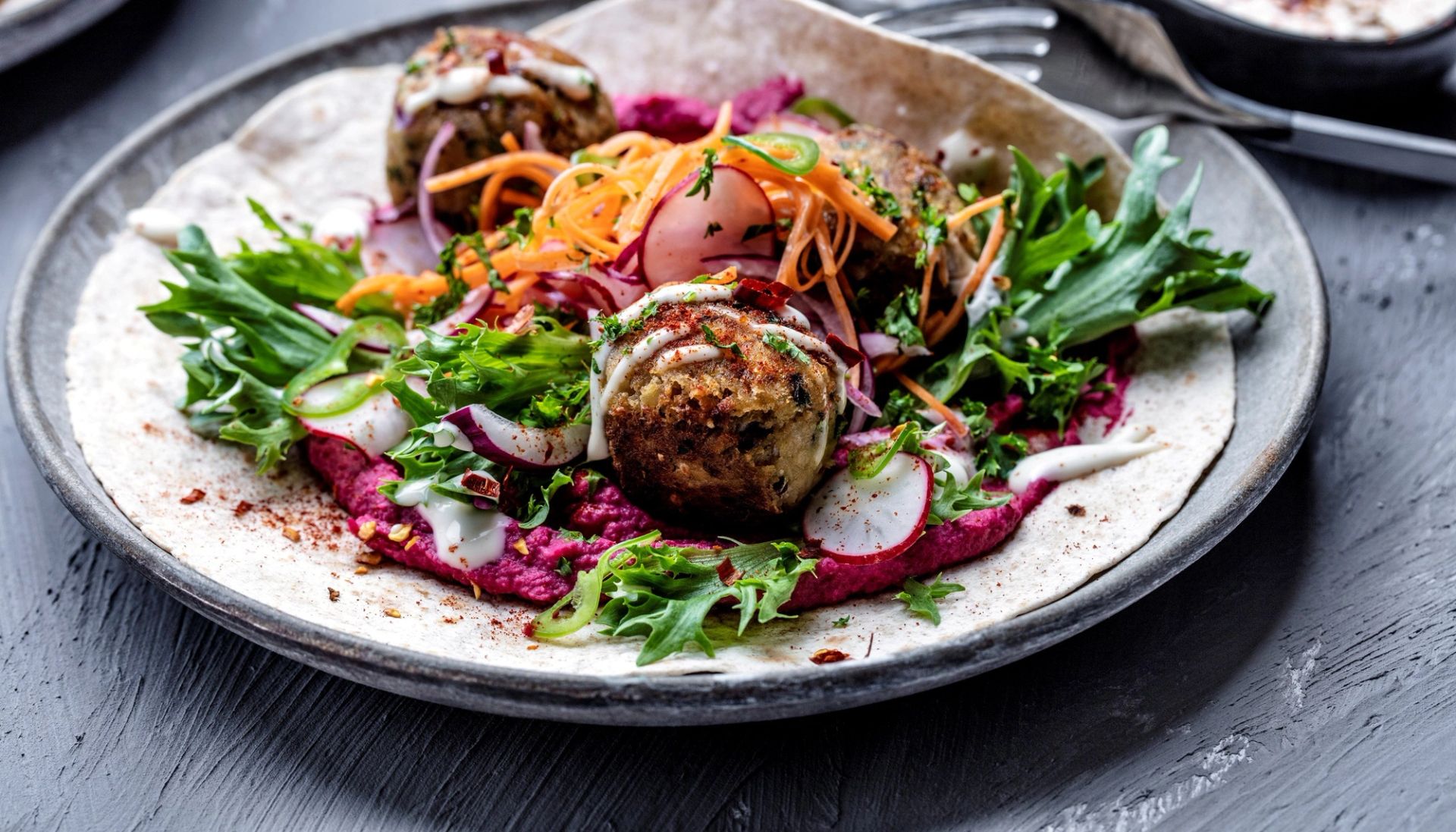 Grov tortillawrap fylt med sprø falafel, kikertboller, rødbethummus og marinert reddik, gulrot og rødløk på mørk tallerken