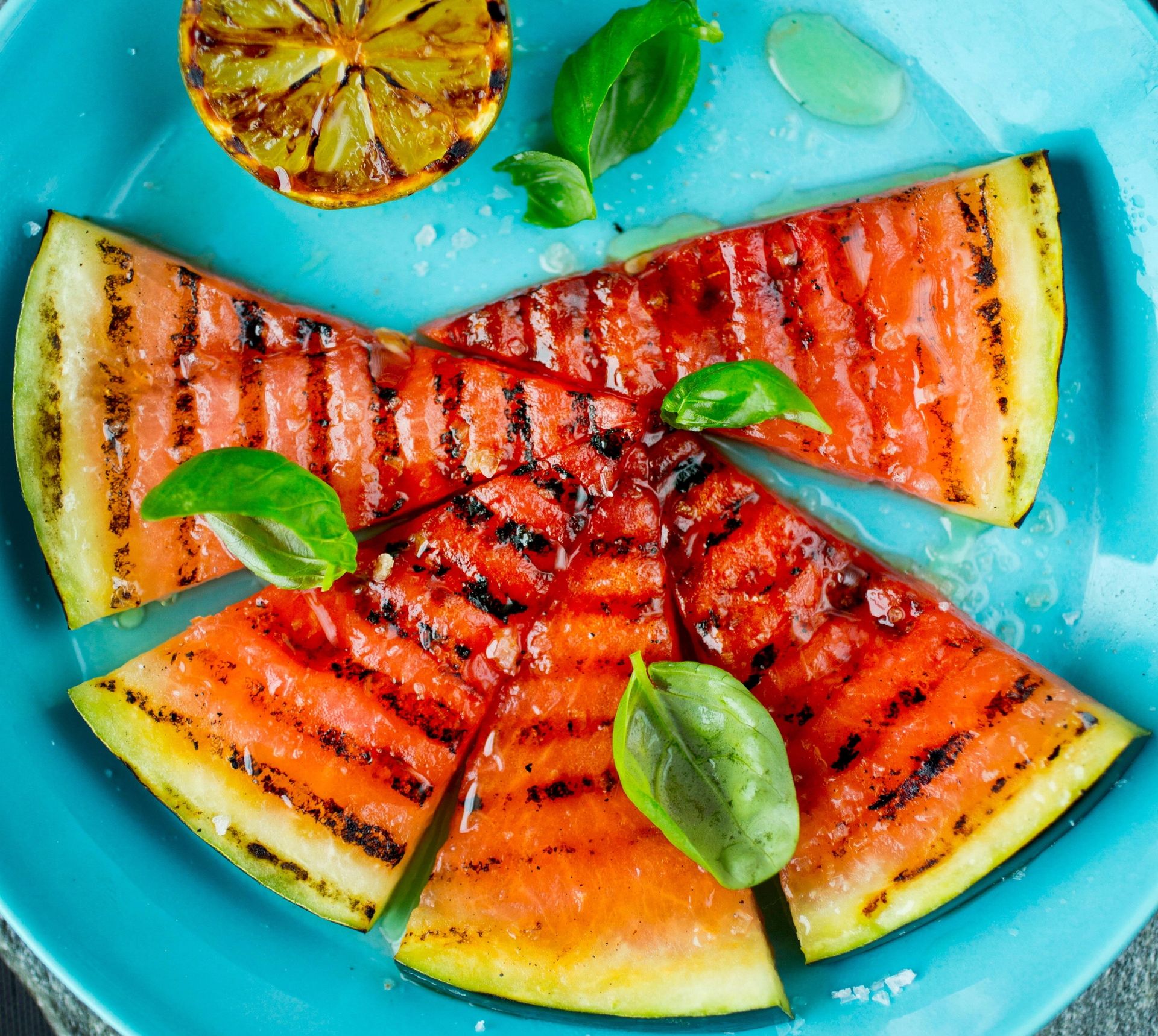 Grillede skiver av vannmelon arrangert i sirkel på en turkis tallerken, toppet med friske basilikumblader og flaksalt, med en grillet halv lime øverst.