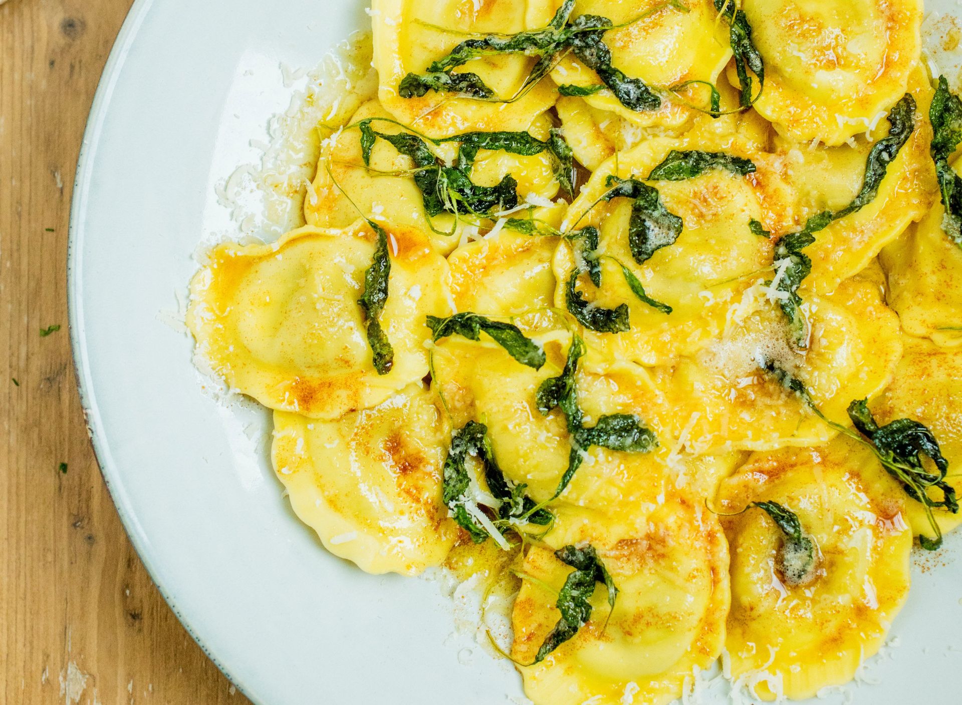 Nærbilde av ravioli i en kremet, gyllen saus toppet med revet parmesan og strimler av spinat, servert på en lys tallerken på et trebord.