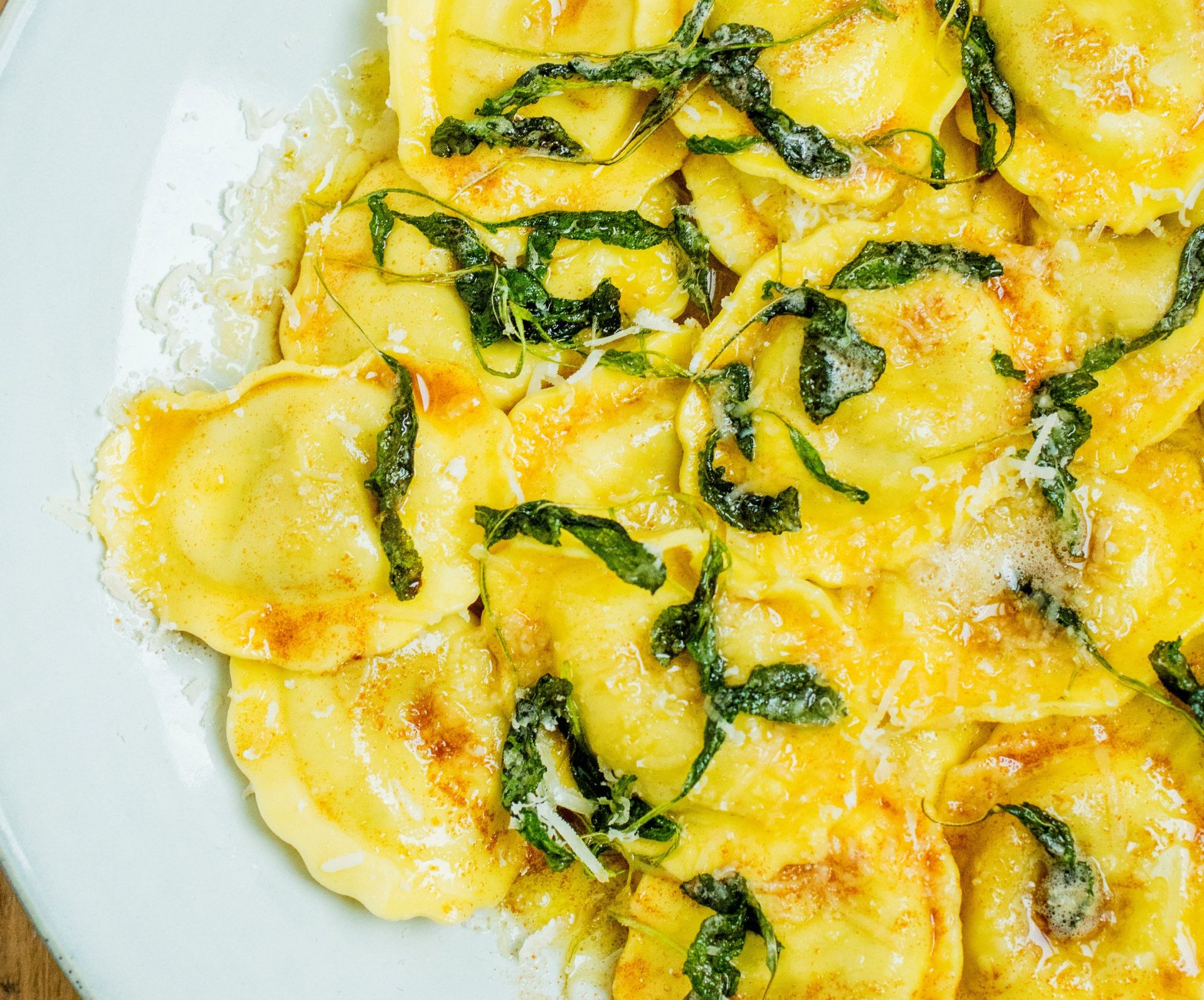 Nærbilde av ravioli i en kremet, gyllen saus toppet med revet parmesan og strimler av spinat, servert på en lys tallerken på et trebord.