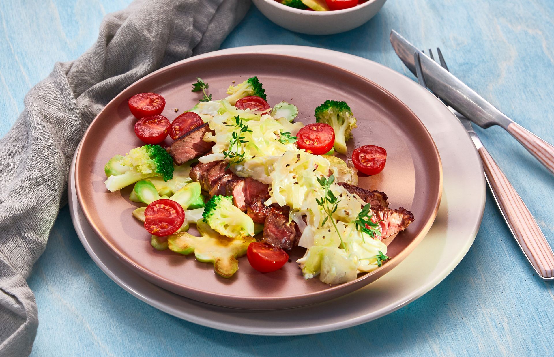 Stekt entrecote servert med kremet kål, brokkolibuketter og halve cherrytomater, garnert med friske urter på en rosa tallerken.