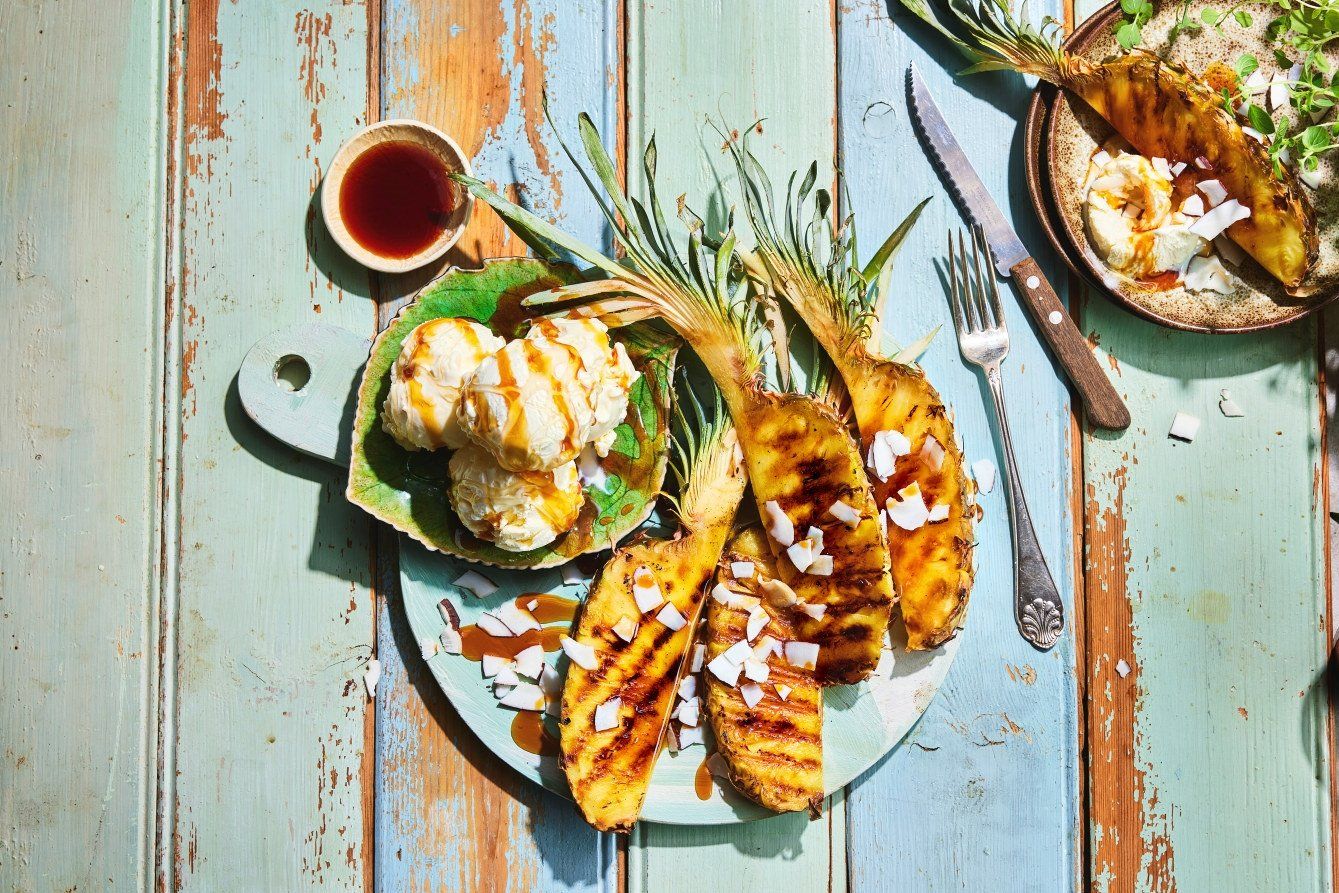 Tre grillede ananasbåter med tydelige grillstriper servert på en lyseblå trefjøl sammen med tre kuler vaniljeis. Desserten er toppet med mørk karamellsaus og hvite kokosflak, og er dander på et rustikt trebord ved siden av en liten skål med saus, en gaffel og en kniv i sterkt sollys.