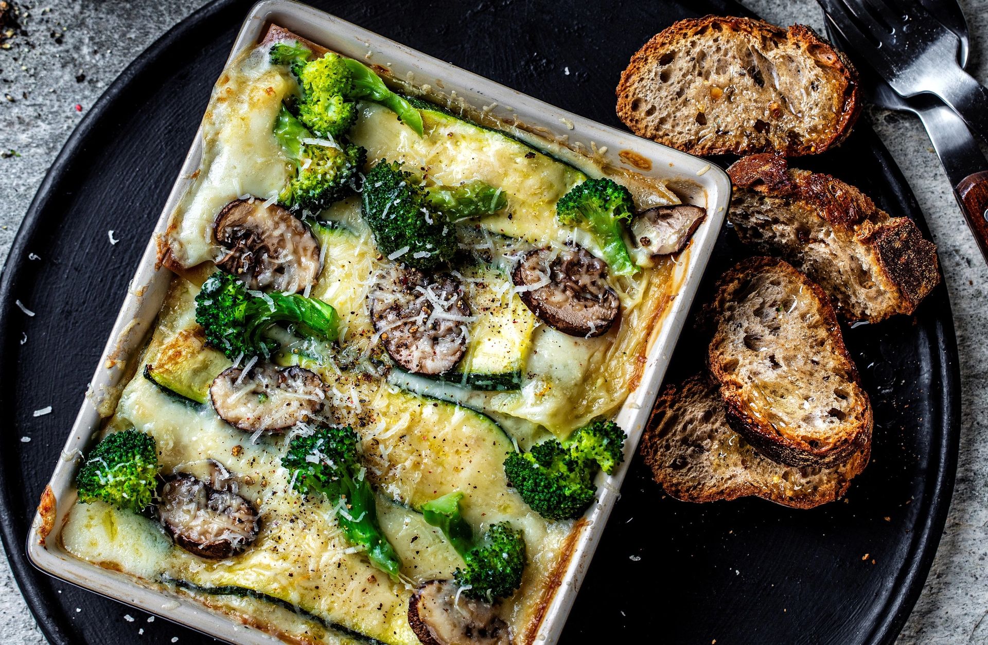 Form med grønn lasagne laget med brokkoli, squash og sopp, toppet med revet ost, servert på et rundt svart fat med ristede brødskiver og parmesan ved siden av.
