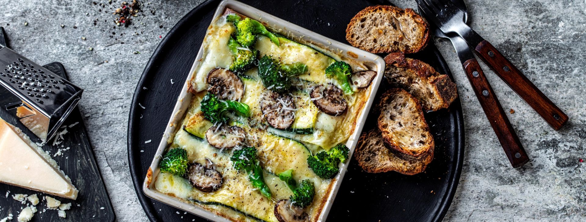Form med grønn lasagne laget med brokkoli, squash og sopp, toppet med revet ost, servert på et rundt svart fat med ristede brødskiver og parmesan ved siden av.