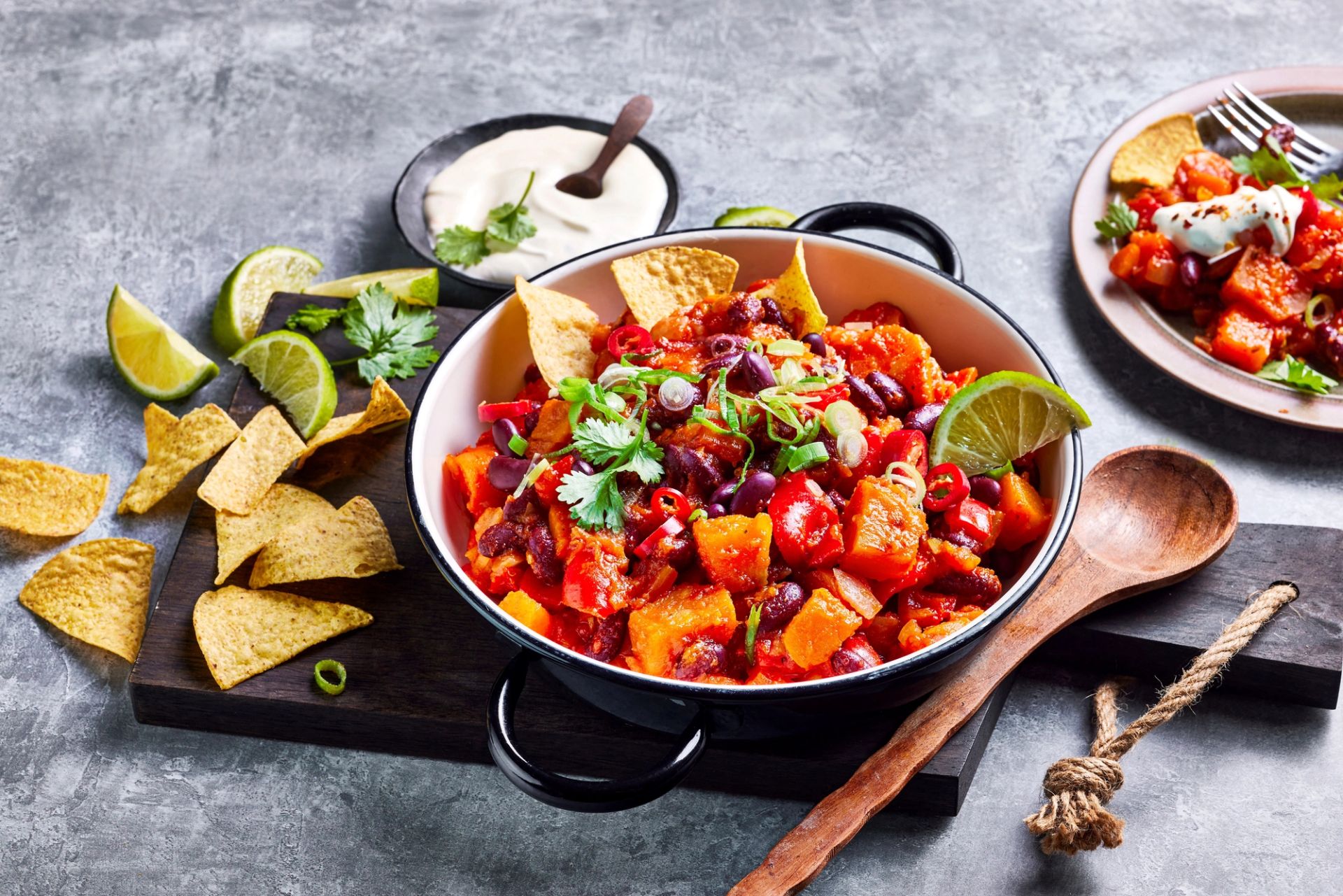 Gryte med søtpotet‑ og bønnegryte i tomatsaus, toppet med tortillachips, frisk koriander og skivet chili. Servert med lime, rømme og nachochips på grå bakgrunn.