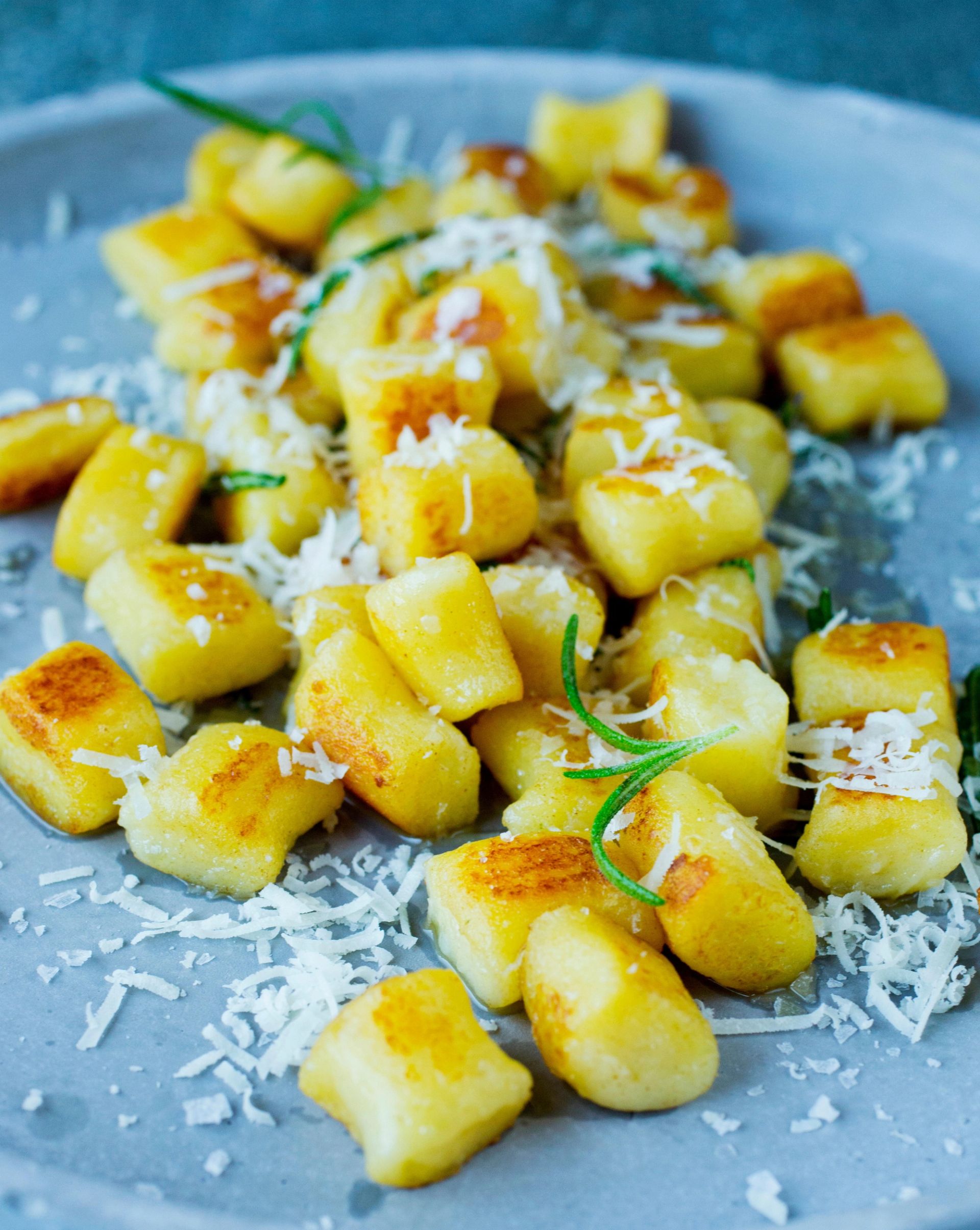Gylne, stekte gnocchi‑biter toppet med revet ost og friske urter, servert på en lys grå tallerken.