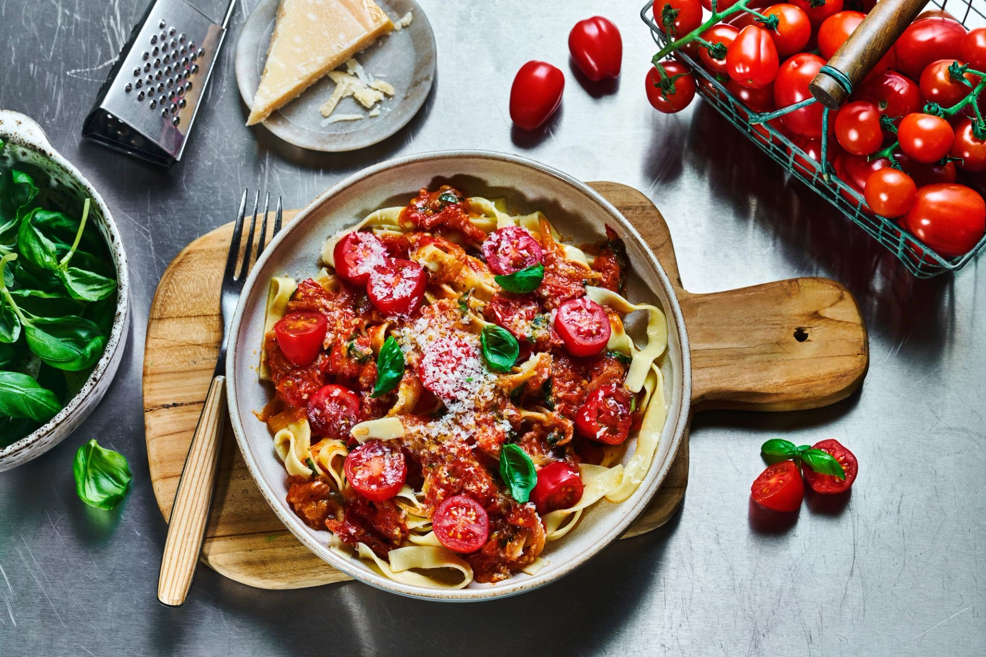 En skål med fersk pasta toppet med tomatbasert saus, cherrytomater, revet parmesan og basilikumblader, plassert på et trebord med parmesan, tomater og frisk basilikum rundt.
