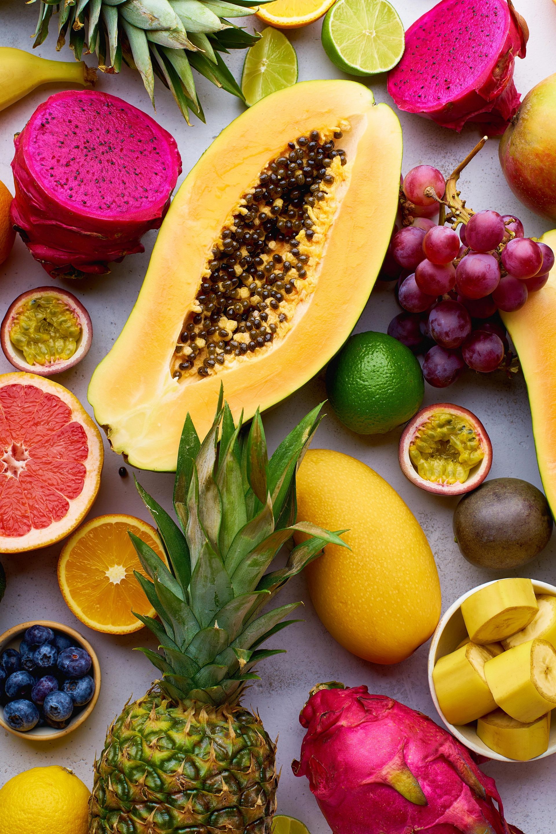 Fargerik samling av tropiske frukter sett ovenfra, inkludert papaya, ananas, mango, dragefrukt, druer, lime, appelsin, grapefrukt og pasjonsfrukt, arrangert på en lys bakgrunn.