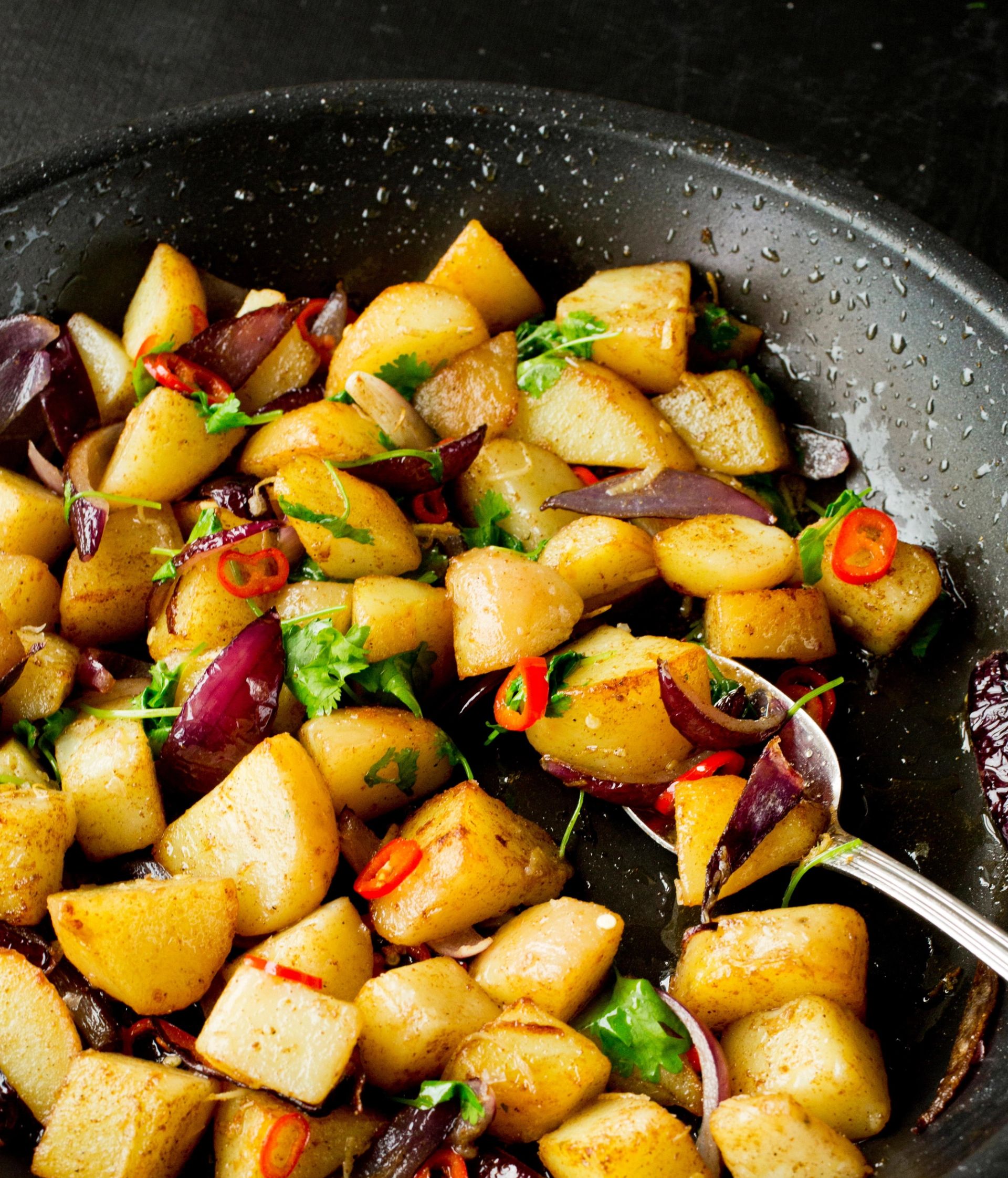 Stekte terninger av pimpernel‑potet i en stekepanne, blandet med rødløk, chili, urter og olje. Potetbitene er gylne og sprø i kantene, og en skje ligger i pannen.