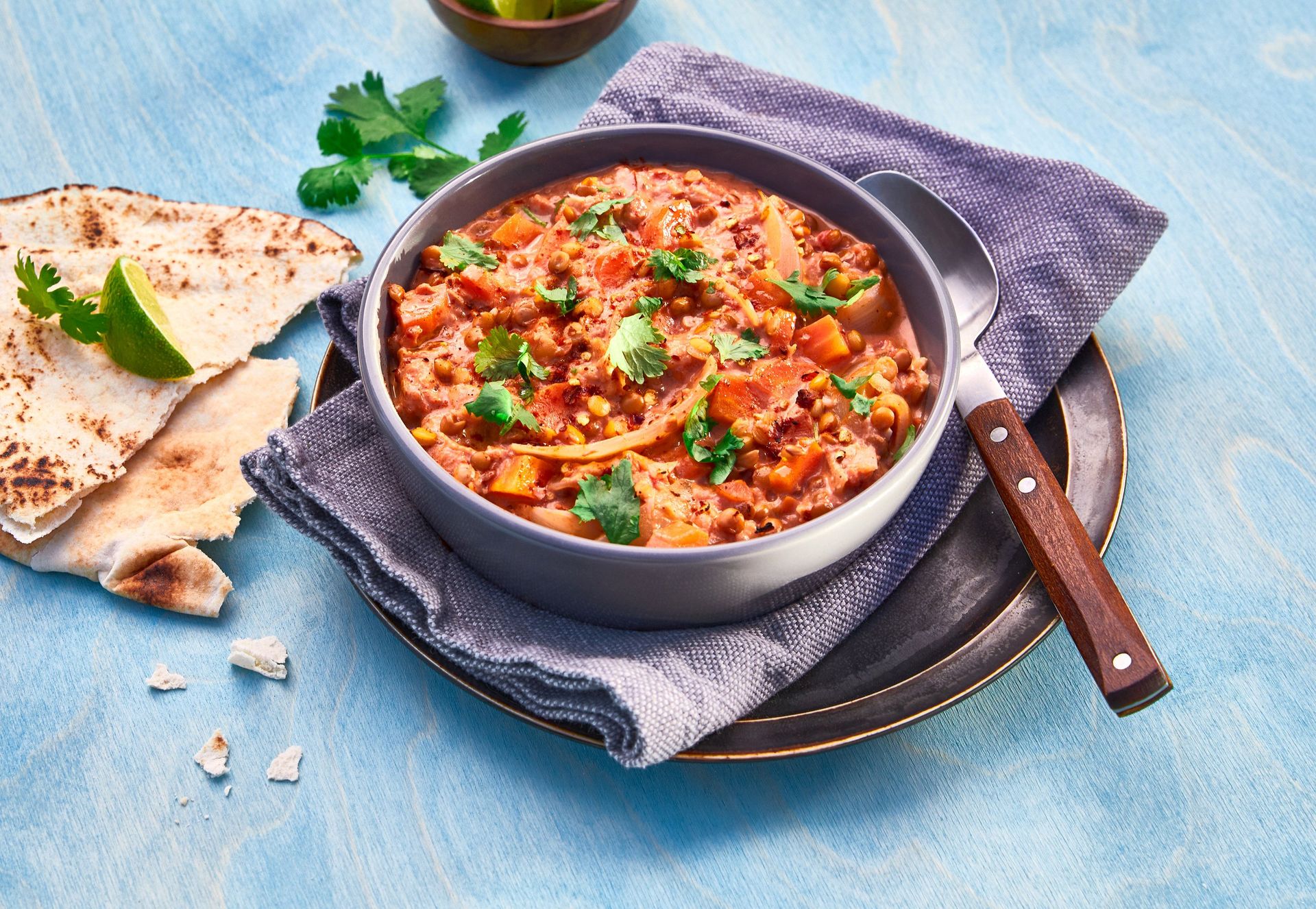 Skål med fyldig dhal laget av linser, gulrøtter, blomkål og gul løk, toppet med koriander, servert med naanbrød og en limebåt på et blått bord.