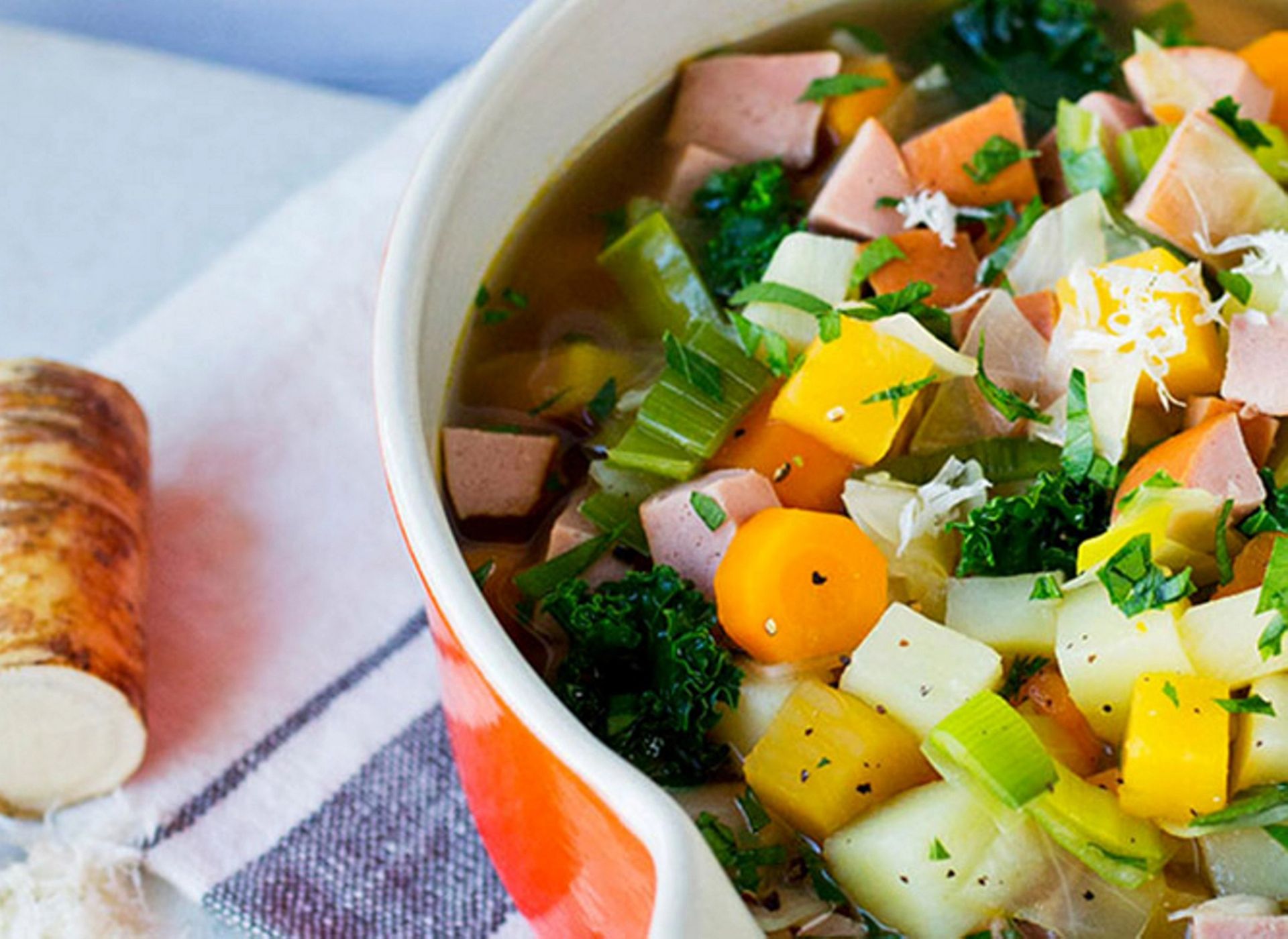 Et nærbilde av en fargerik og mettende grønnsakssuppe servert i en oransje gryte. Suppen inneholder biter av pølse, gulrot, kålrot, potet og grønnkål i en klar kraft.