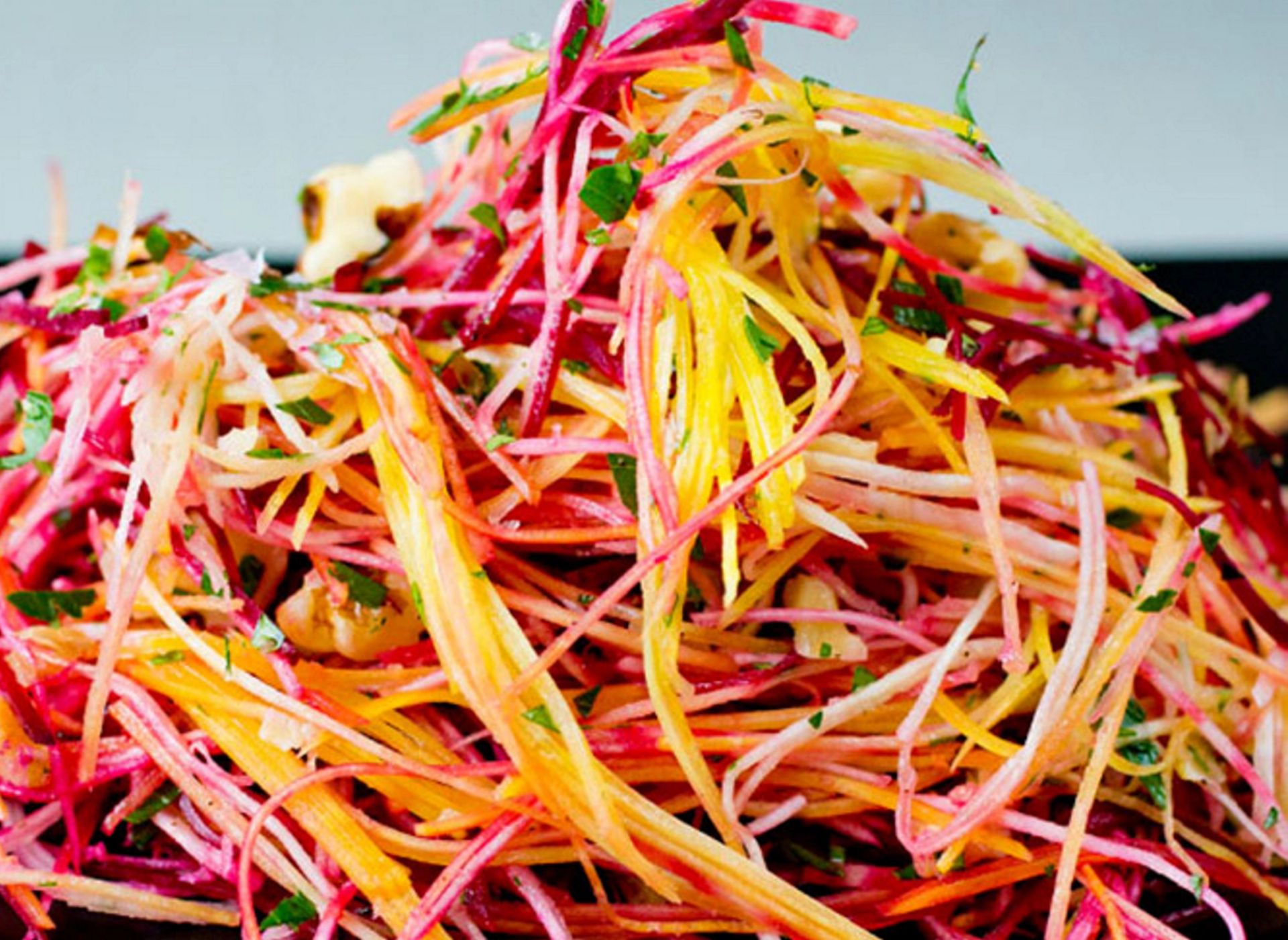 En fargerik råkostsalat bestående av tynne strimler av rotgrønnsaker i nyanser av lilla, gult og oransje. Salaten er blandet med hakkede valnøtter og friske grønne urter.
