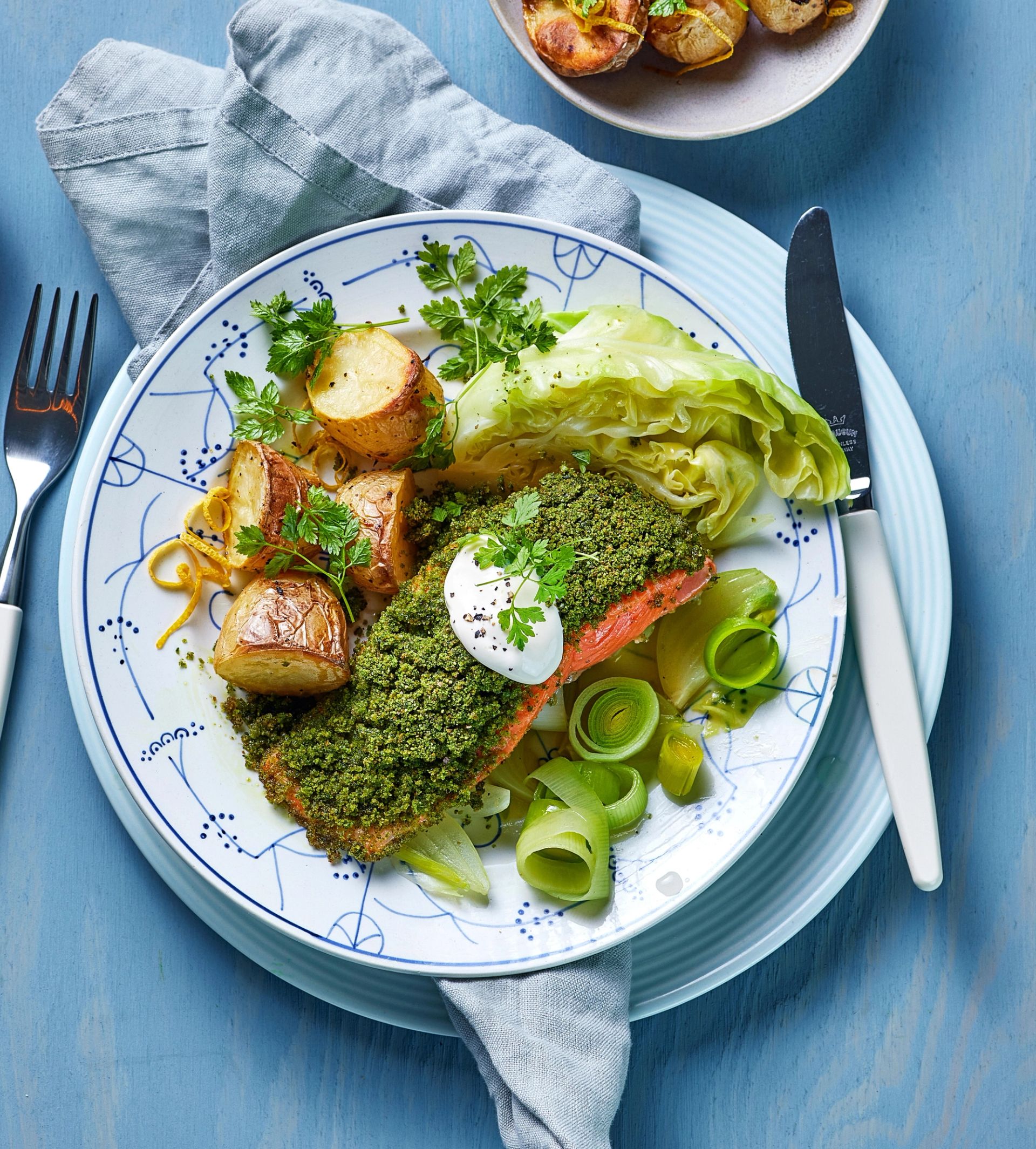 En tallerken med et ferdig måltid servert på et blått bord. Midt på tallerkenen ligger et stykke fisk med grønn urteskorpe, toppet med en liten klatt saus. Rundt fisken ligger flere tilbehør: ovnsbakte små poteter med gyllen overflate, dampet nykål i store, lysegrønne blader, og skiver av purre rullet i små spiraler. Retten er pyntet med ferske urter. Tallerkenen har et hvitt og blått mønster og står på et brettet, lyst serviett. Ved siden av ligger en kniv, og øverst i bildet er en bolle med flere poteter delvis synlig. Lyset er jevnt og fremhever de friske fargene i retten.