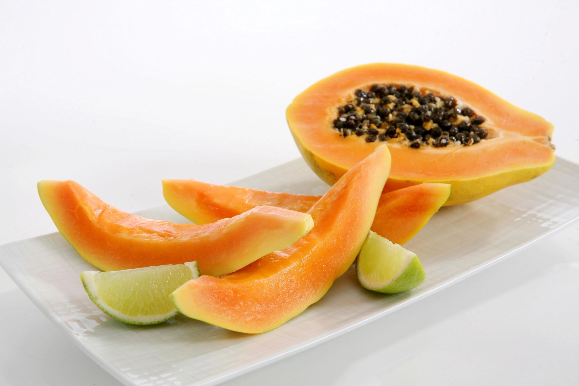 Skiver av moden papaya servert på et hvitt fat, med halvert papaya med frø i bakgrunnen og limebåter ved siden av.