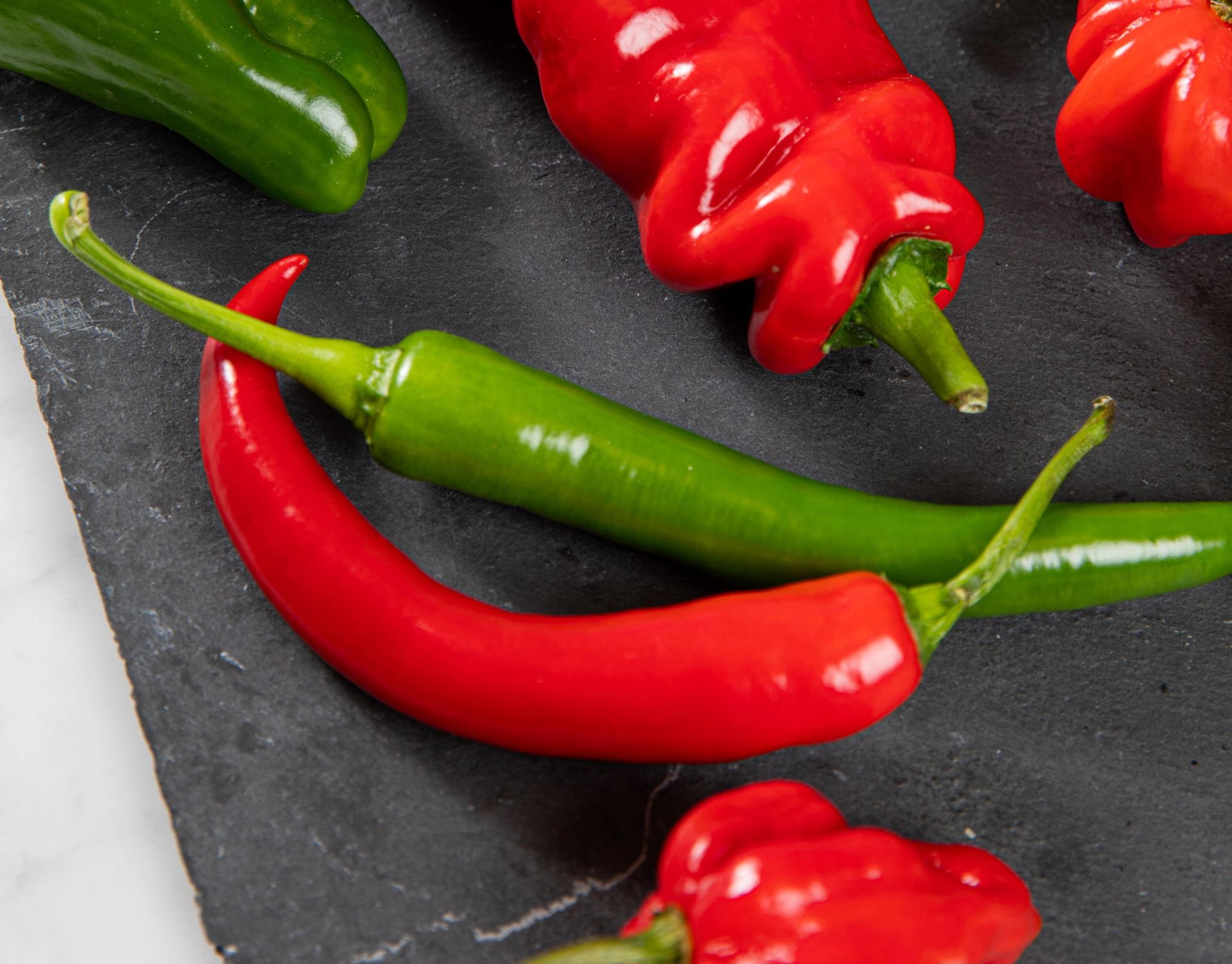 Flere røde og grønne chilipepper ligger sammen på en mørk overflate.
