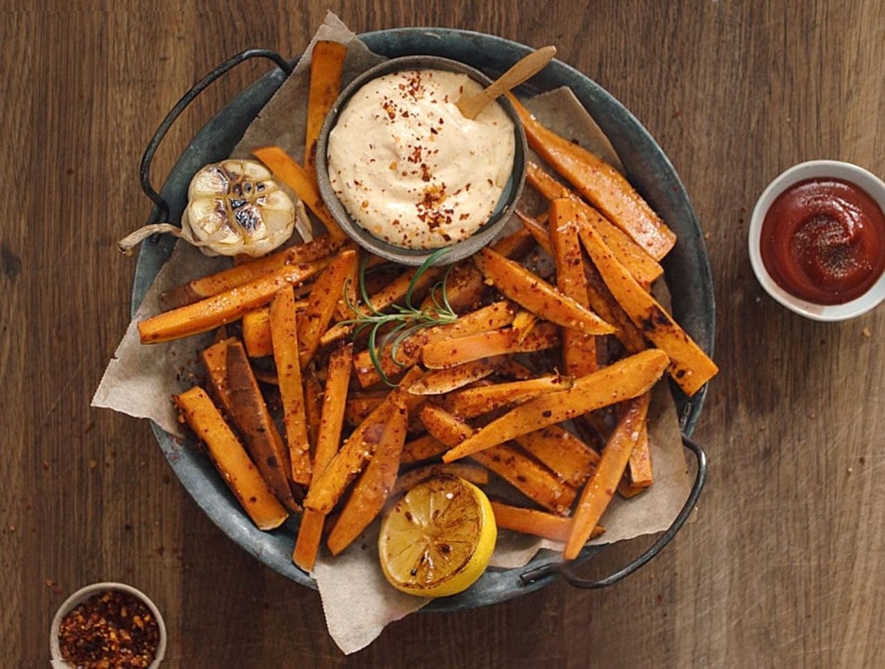 Ovnsbakte søtpotetfries servert i et rustikt fat med kremet dip i midten. Fatet er pyntet med grillet sitron, bakt hvitløk og frisk rosmarin, og omgitt av små skåler med ketchup og chiliflak på et mørkt trebord.