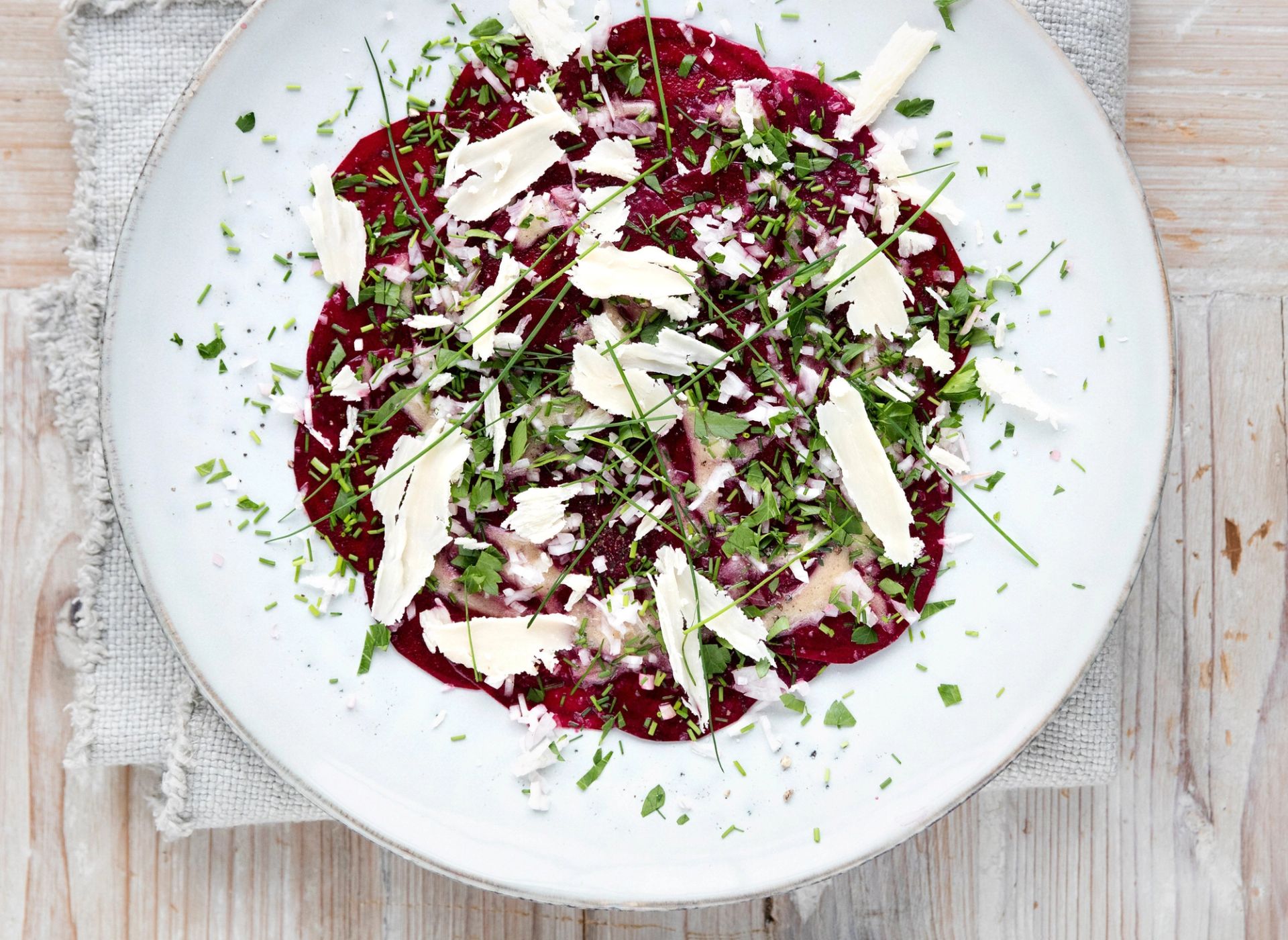 Tallerken med tynne skiver rødbet arrangert som carpaccio, toppet med finhakket rødløk, urter og store flak av parmasan, sett ovenfra på et lyst trebord.