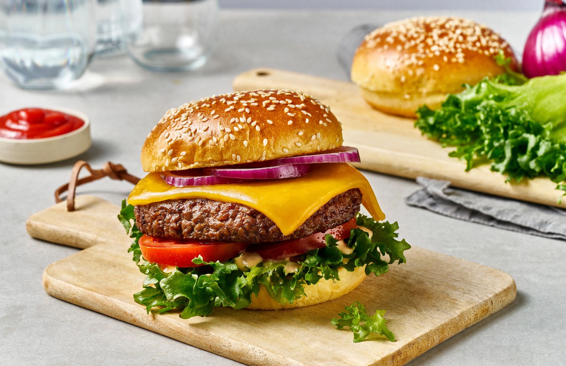 Burger med sesambrød fylt med saftig kjøttburger, smeltet gulost, skiver av tomat og rødløk, sprø salat og dressing, servert på en trefjøl med ketchup og ekstra tilbehør i bakgrunnen.