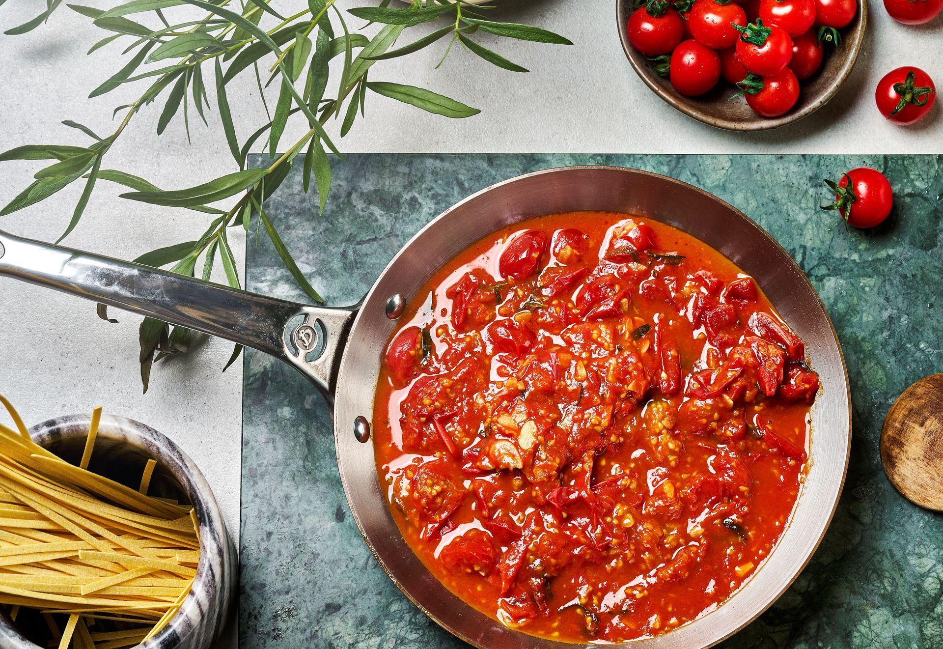 Nærbilde av en tomatsaus med hele og oppdelte cherrytomater i en panne, fotografert ovenfra. I bakgrunnen ligger tørket pasta i en bolle, ferske cherrytomater og friske urter på et kjøkkenbenk.
