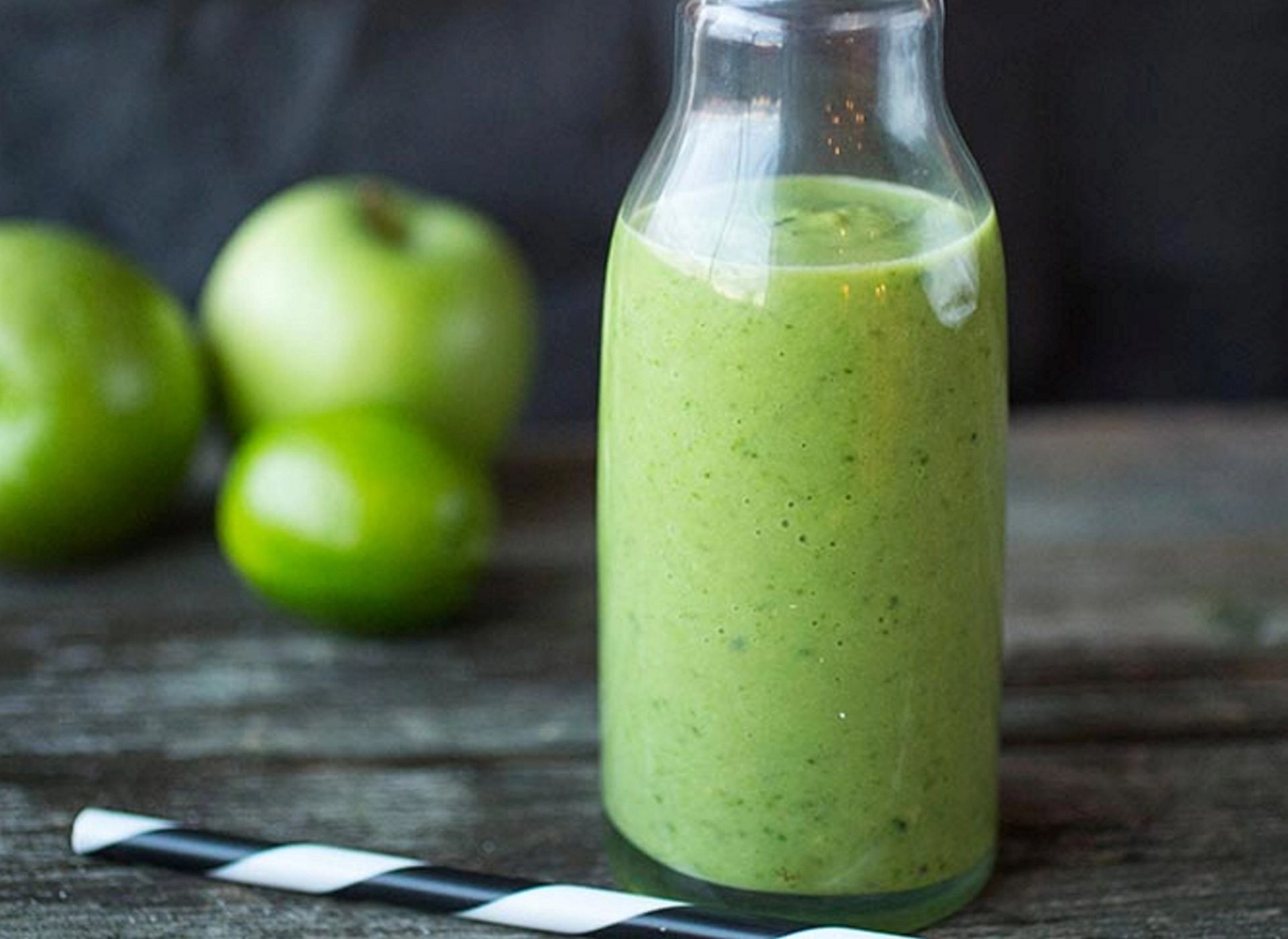 Kremet grønn supersmoothie i glassflaske, med epler og sitrus i bakgrunnen.