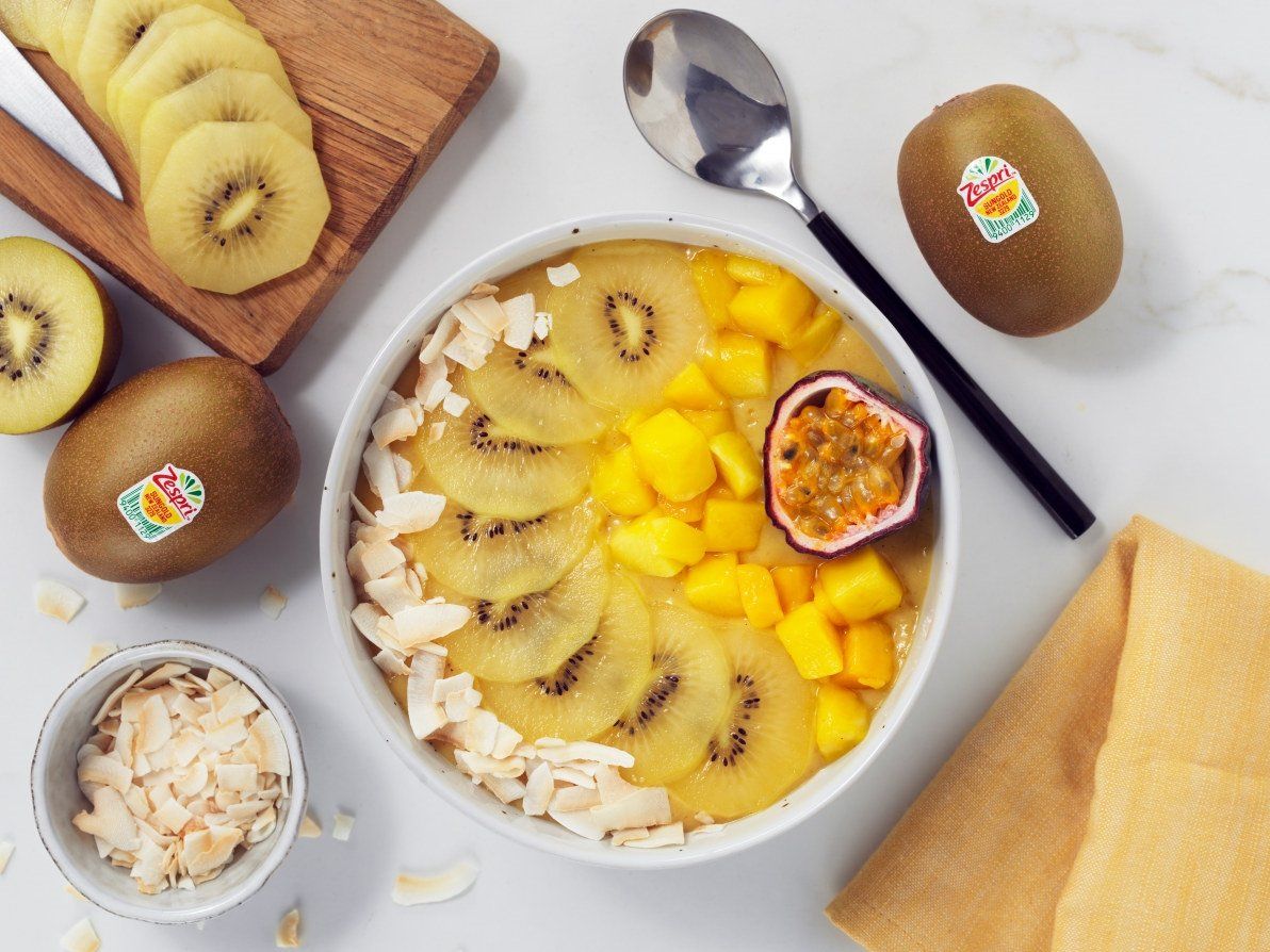 En frisk smoothie-bolle toppet med skiver av gul kiwi, terninger av mango og en halv pasjonsfrukt. Rundt bollen ligger hele og skivede kiwier, en liten skål med kokosflak, en skje og en gul serviett på en lys overflate.