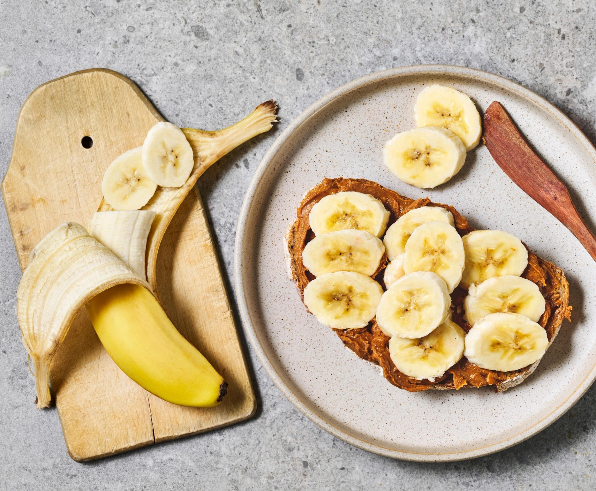 Nærbilde av en halv-skrelt banan og en brødskive med peanøttsmør og skivede bananer på toppen.