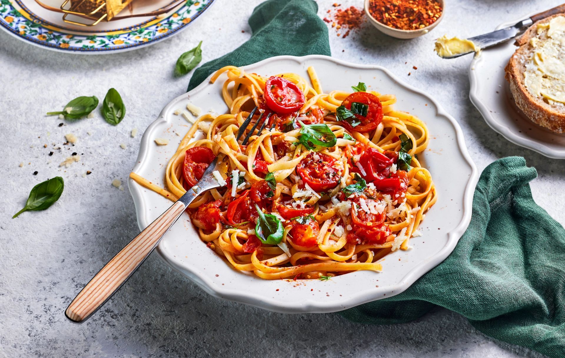 En dyp, hvit porselenstallerken fylt med rykende fersk spaghetti i en fyldig tomatsaus. Pastaen er toppet med saftige, bakte cherrytomater, revet parmesan og friske basilikumblader.