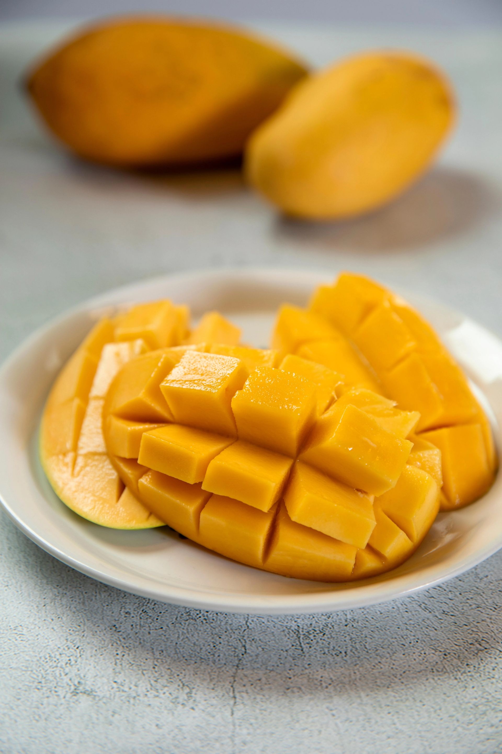 Oppskåret mango i rutenettmønster servert på hvit tallerken, med hele mangoer i bakgrunnen.