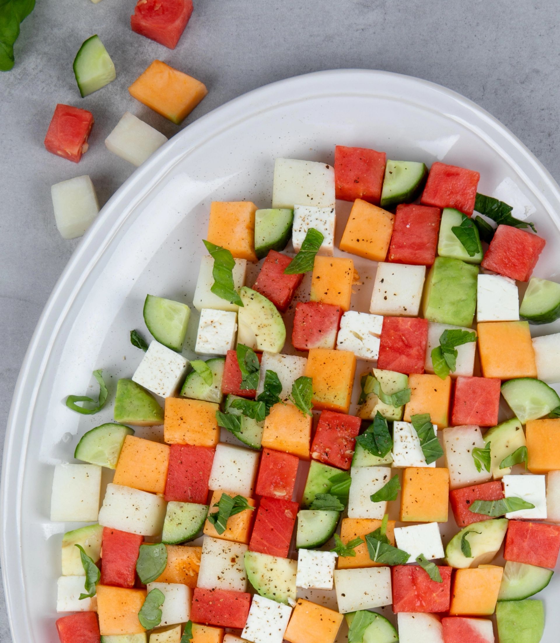 Terninger av melon i røde, grønne, hvite og oransje farger arrangert i et rutemønster på et hvitt fat, toppet med friske mynteblader og krydder, sett ovenfra på en lys bakgrunn.