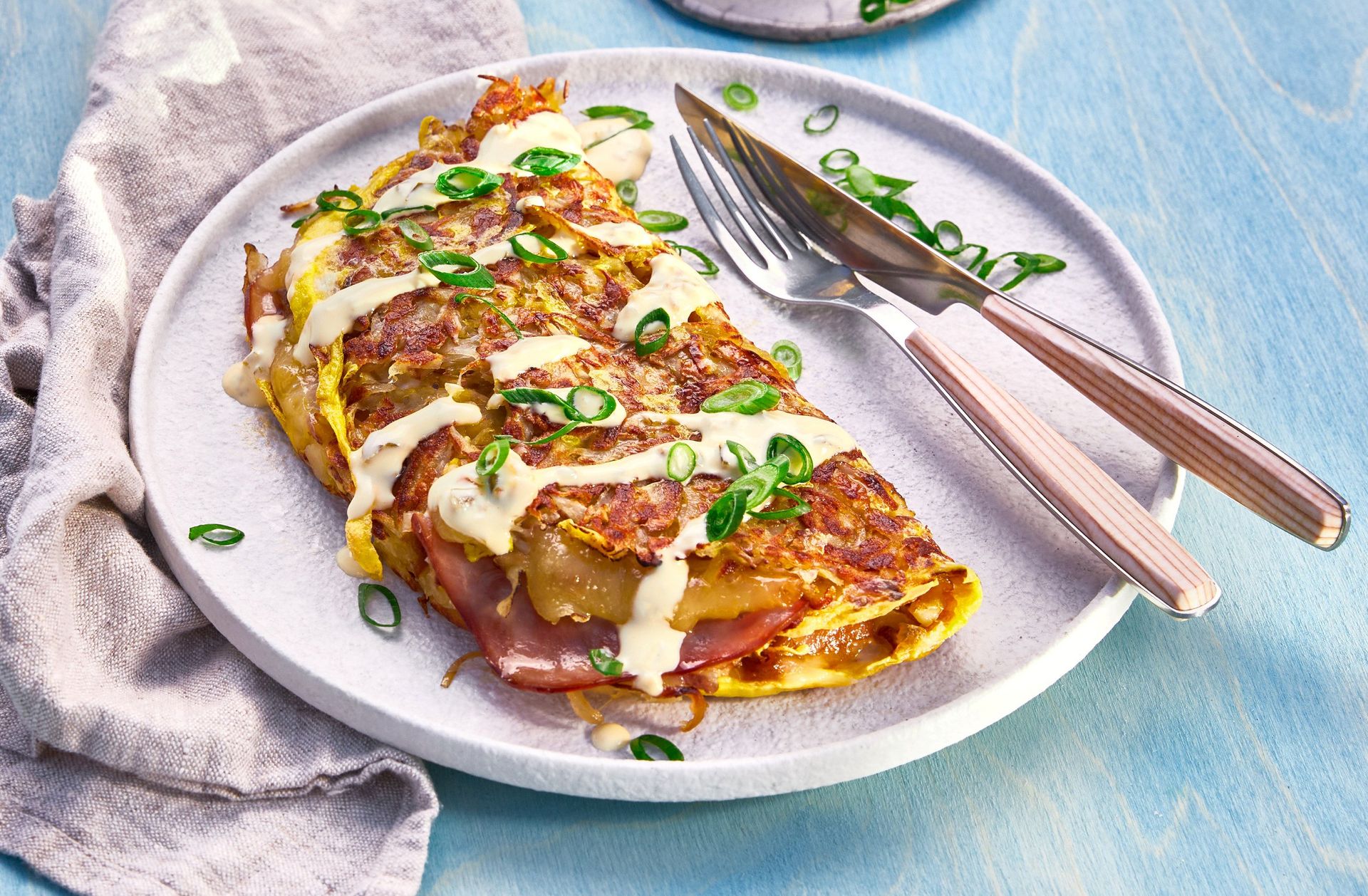 “Okonomiyaki (japansk pannekake) med kål, bacon og vårløk, toppet med saus og kremet dressing, servert på en tallerken.”