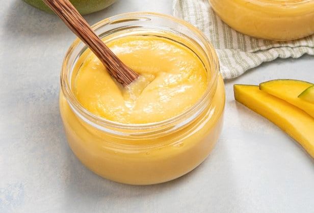 Små glass fylt med kremet, gul mango står på en kjøkkenbenk, sammen med sammen med både hel og skivet mango. En treskje står i et av glassene.