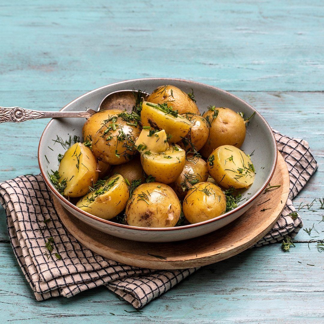 Kokte nye poteter med smør og dill i en hvit skål, servert på et trefat med rutete kjøkkenhåndkle, på et turkist trebord.