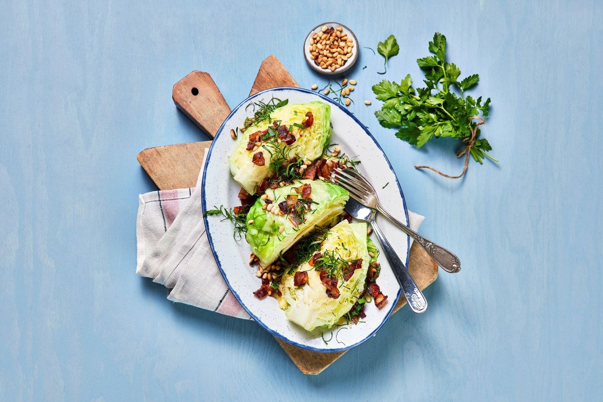 Gratinert nykål med smeltet ost, ristede pinjekjerner og sprøstekt bacon servert på fat.