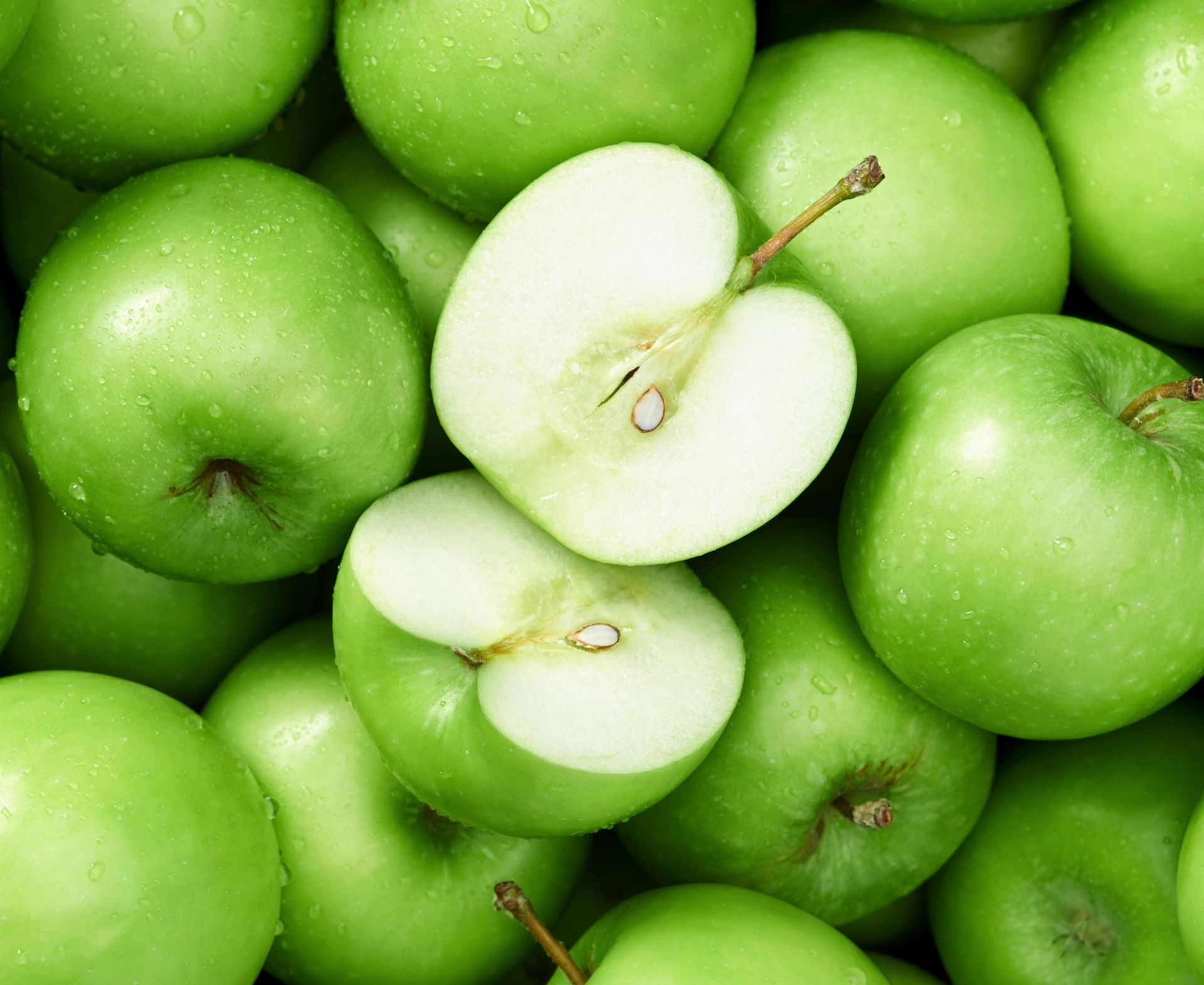 Nærbilde av mange friske, grønne epler som ligger tett sammen og dekker hele bildet. Ett eple er delt i to i midten, slik at den lyse innsiden og kjernehuset med frø er synlig. Eplene har glatt, blank overflate med små naturlige merker og jevn grønn farge.