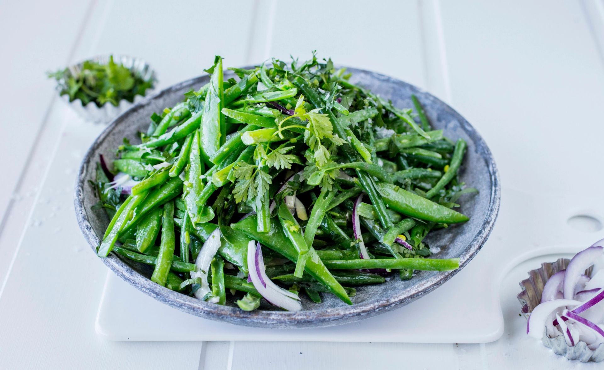 Salat av friske grønne bønner, sukkererter og urter, blandet med tynne skiver rødløk og servert på et grått fat.